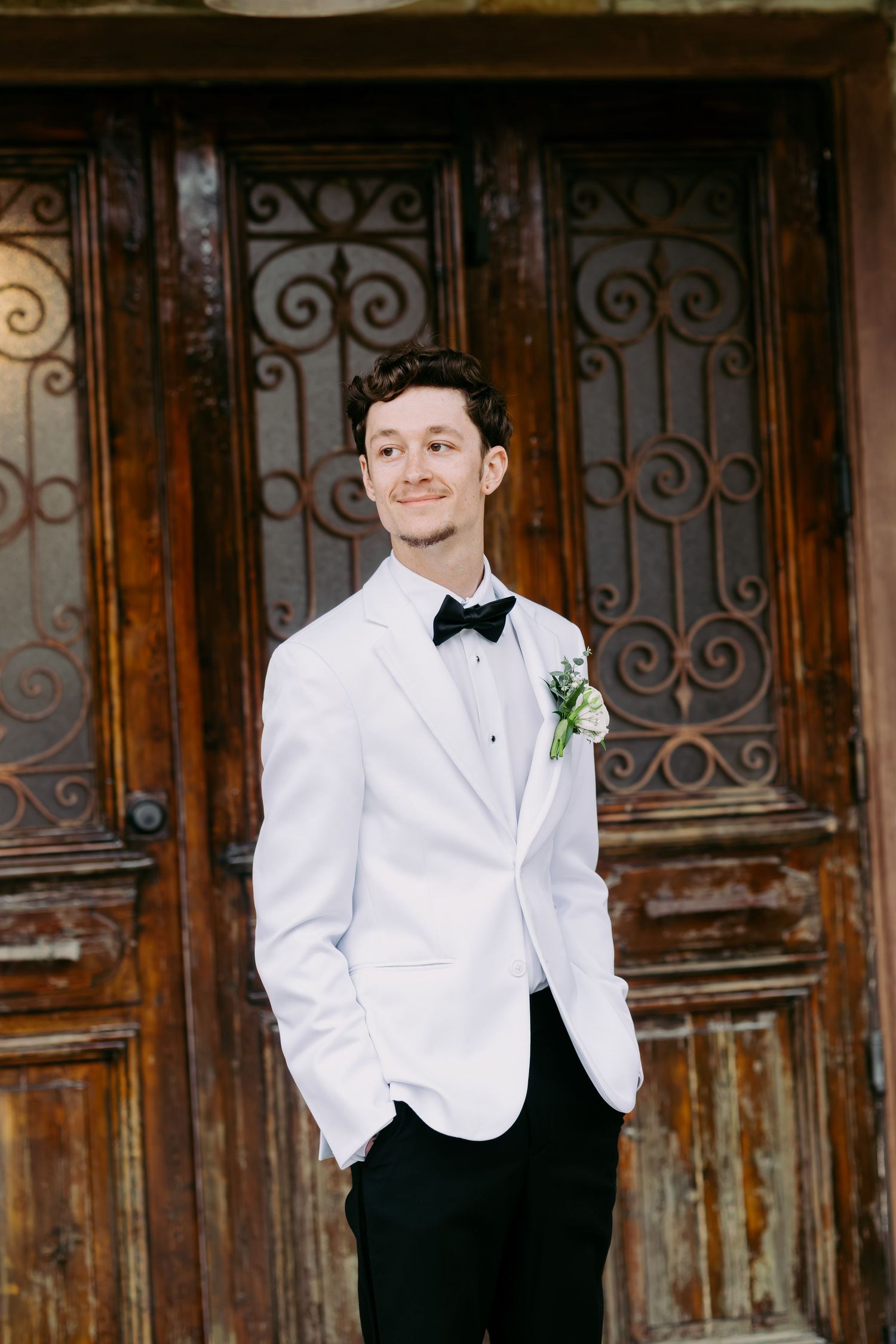 Handsome groom in white tuxedo jacket and black bow tie posing before ornate vintage wooden doors.