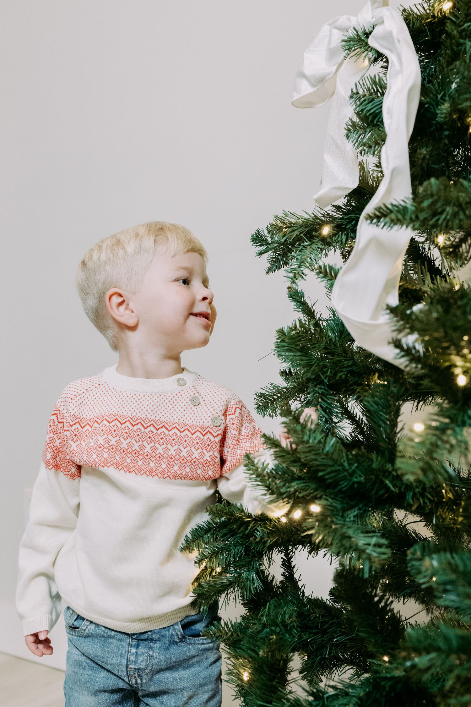 Cute blonde boy in a Nordic sweater looking up at a decorated Christmas tree with white ribbon and lights.