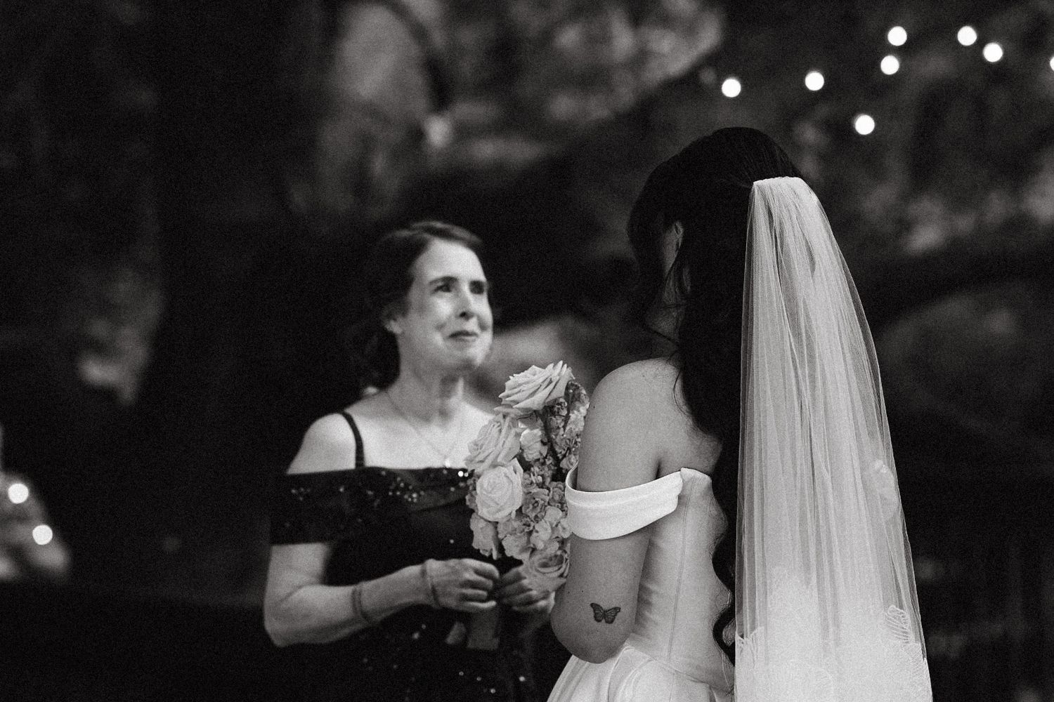 A bride in her wedding dress laughing and holding hands with her mother outdoors.