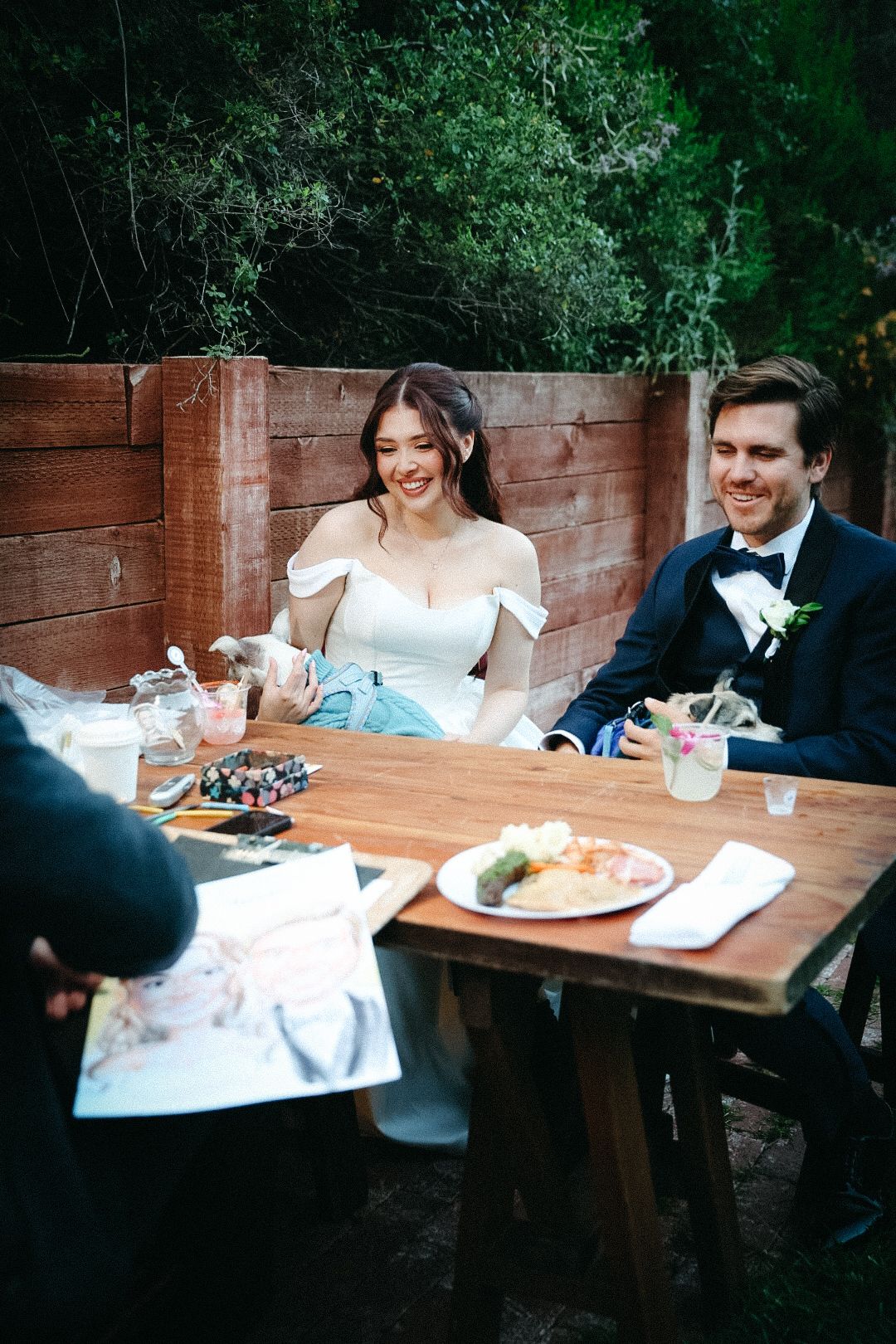 A bride and groom sitting at their wooden reception table smiling and holding their pet dogs.