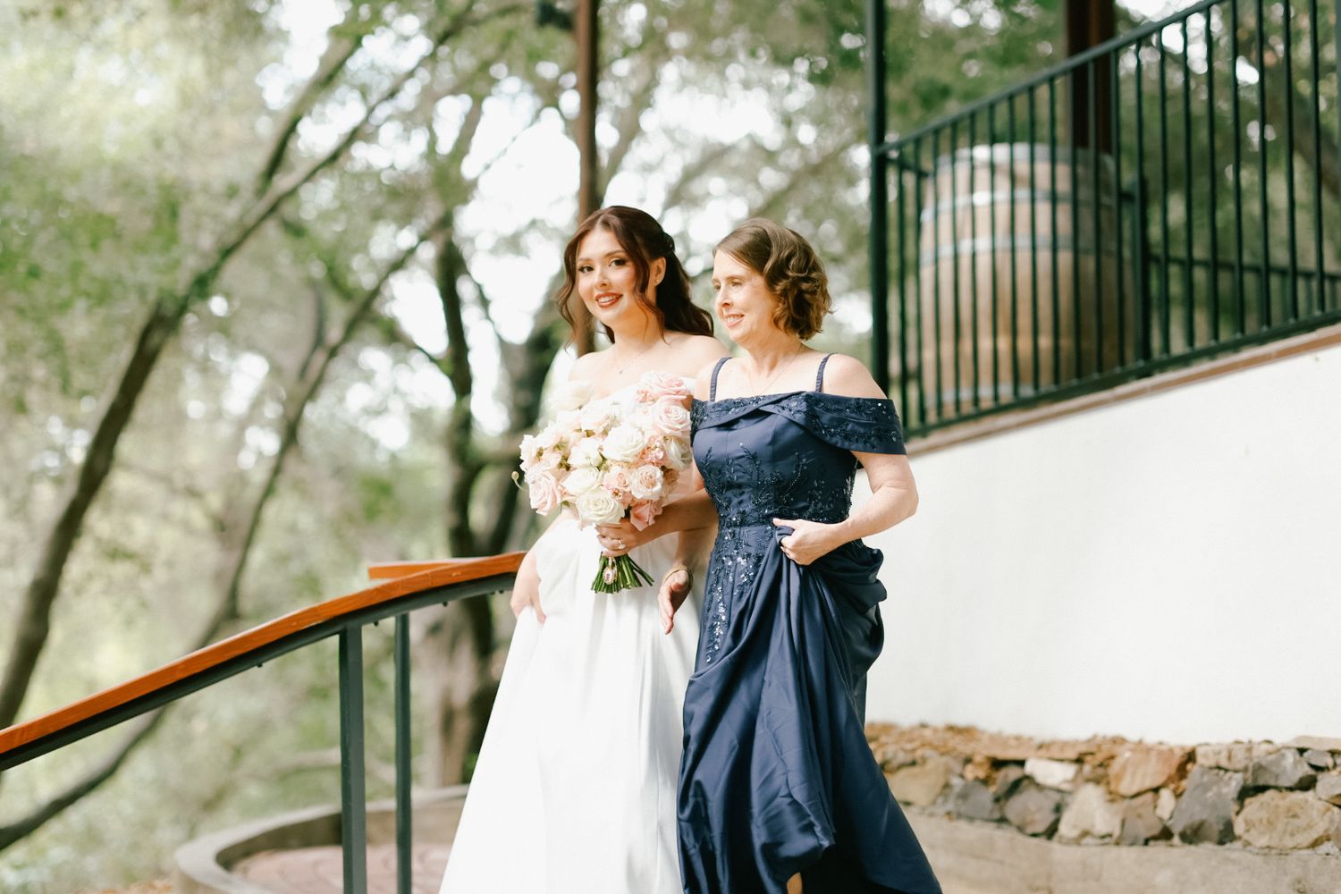 A bride in her wedding dress laughing and holding hands with her mother outdoors.
