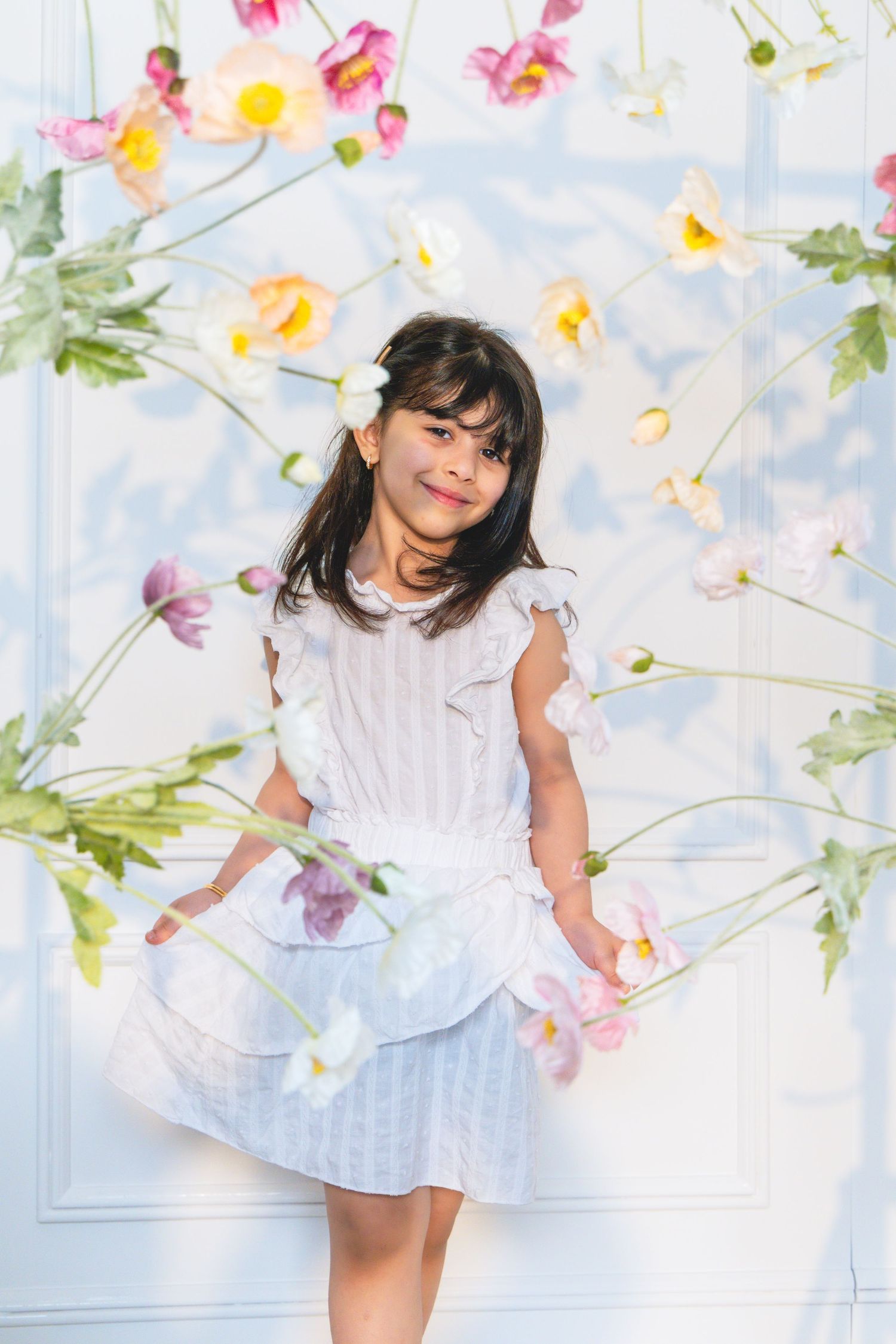 Young girl in white dress posing among hanging pink flowers against a light background.