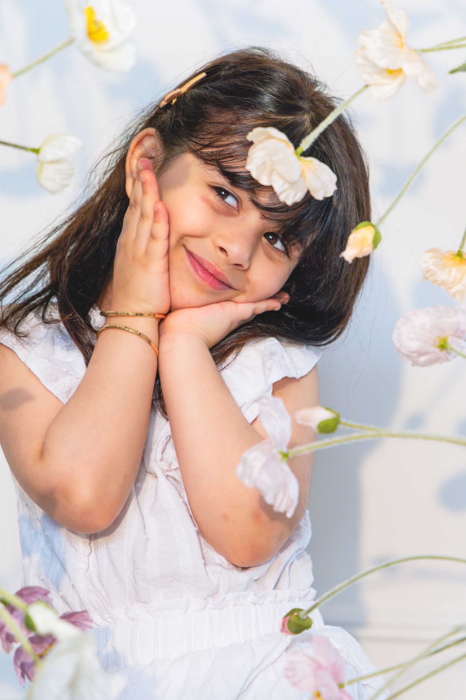 Young girl in white dress with flower in hair, hands on cheeks, smiling among blooming flowers.