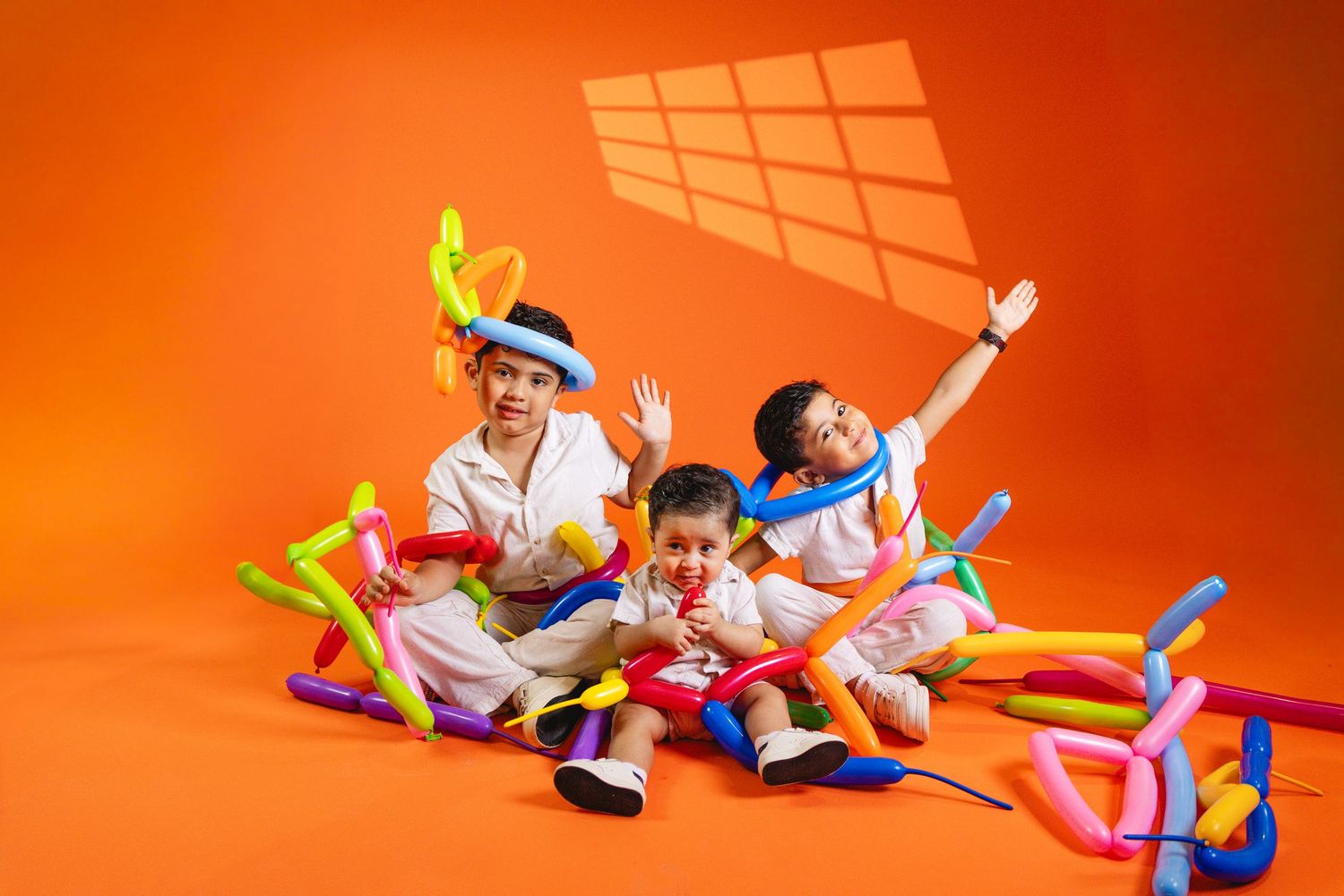 Three playful children surrounded by colorful balloon animals on orange studio background.