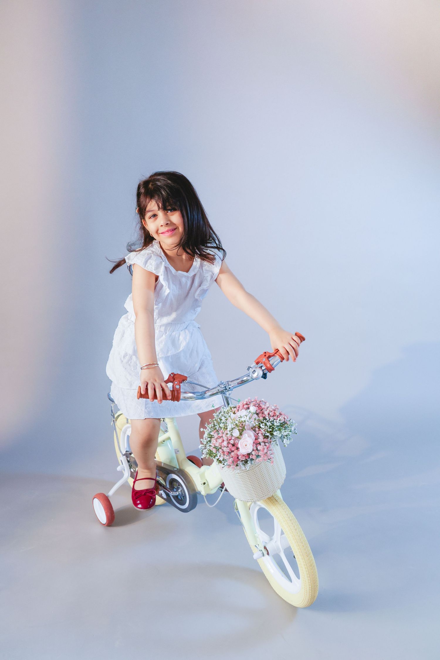 Young girl in white dress smiling on a vintage kids bicycle with flower basket and training wheels.