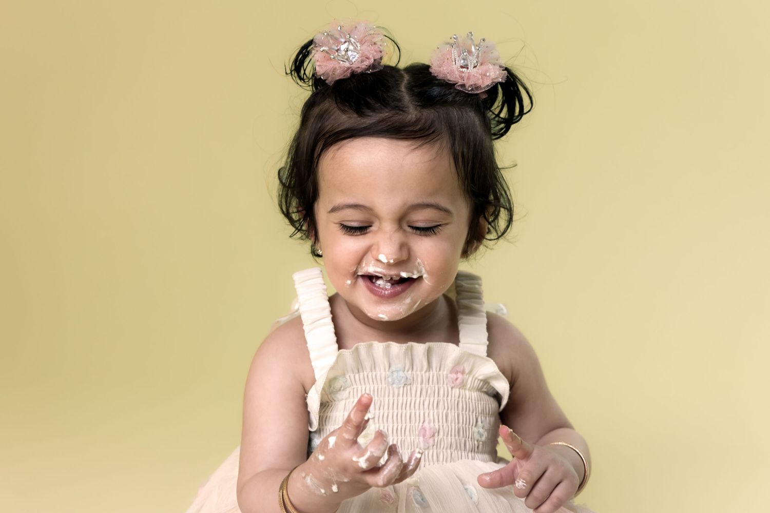 Joyful baby girl with pink bow pigtails laughing during cake smash on yellow background.