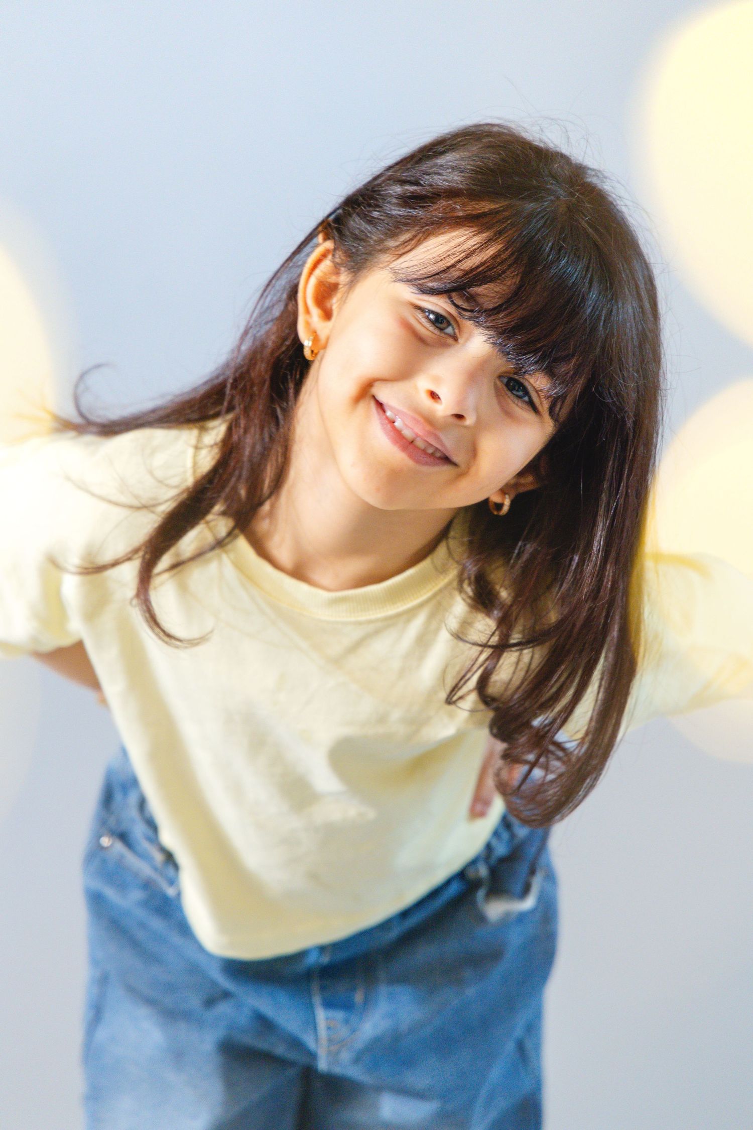 A smiling young girl with long dark hair wearing a yellow t-shirt and jeans poses cheerfully against a light background.