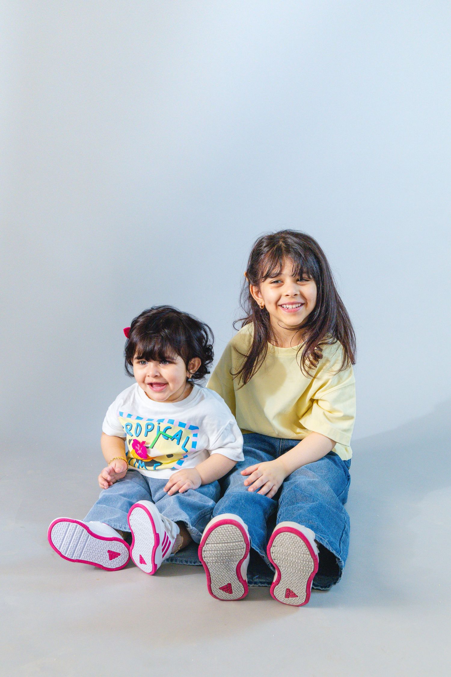 Two young girls sitting together smiling, wearing jeans and pink sneakers, against a light gray background.