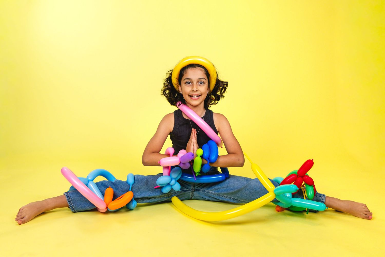Young girl sitting cross-legged on yellow background, smiling, surrounded by colorful twisted balloon animals.