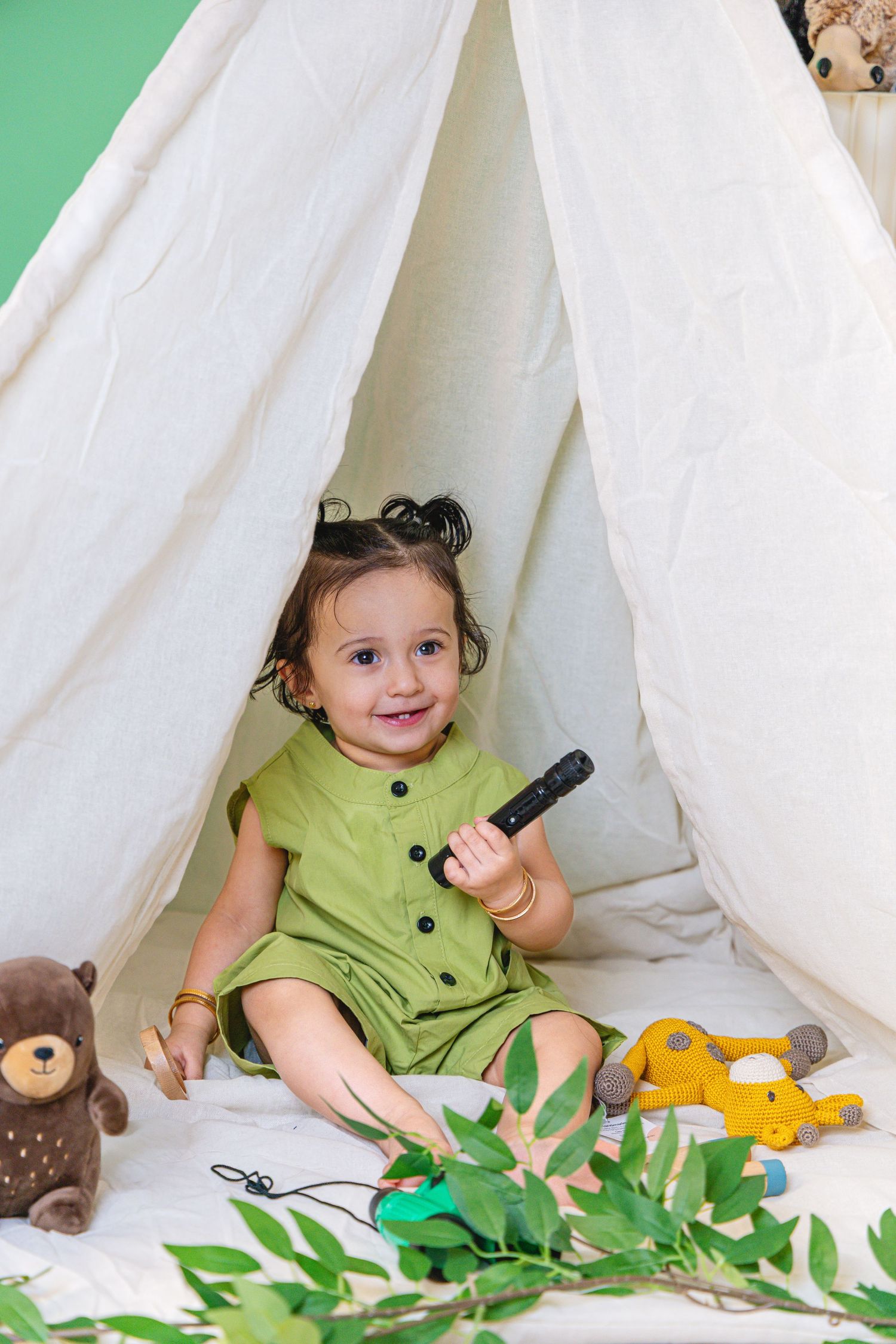 Smiling toddler girl in green dress sitting inside white teepee tent with stuffed animal toys.