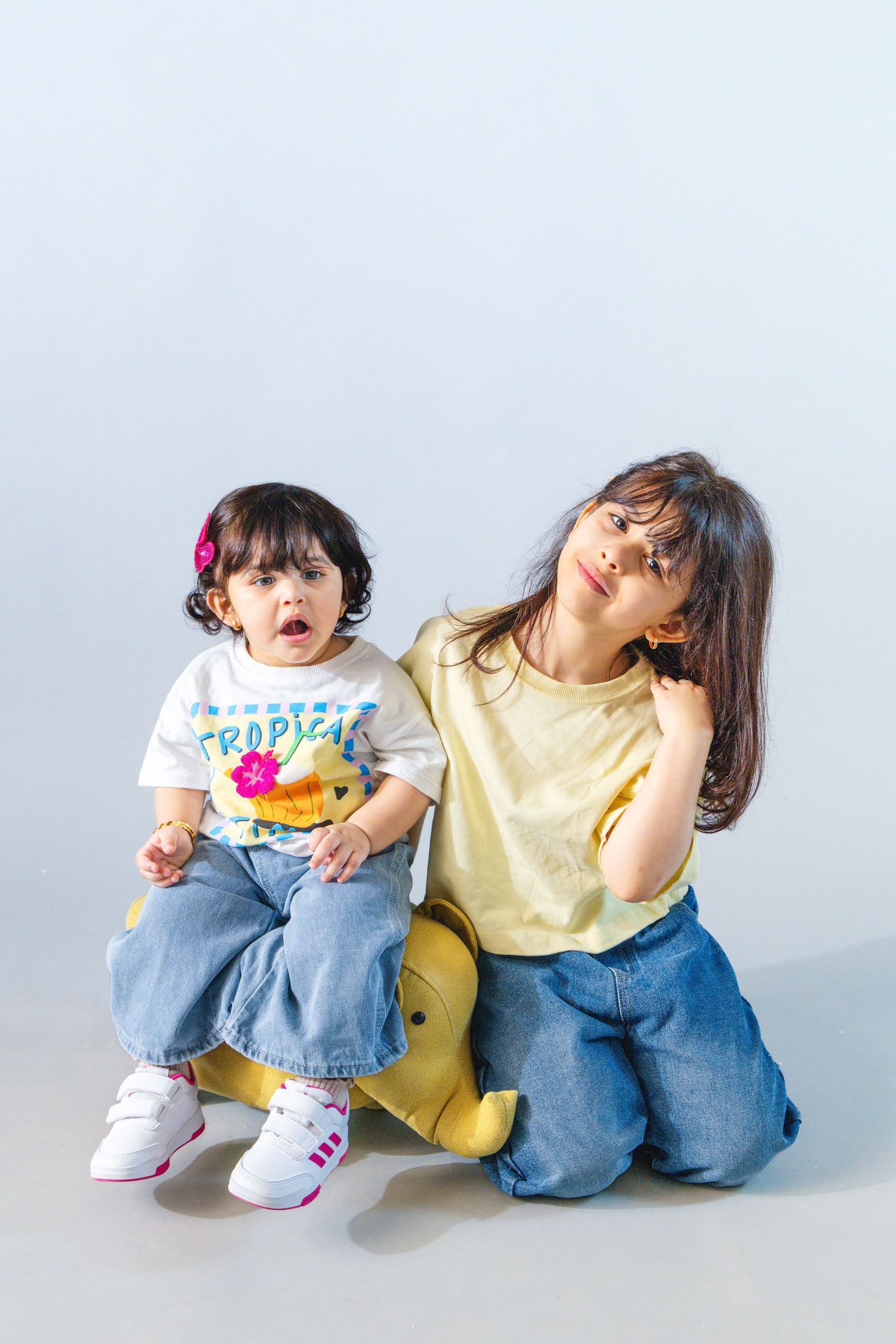 Two young girls in jeans and colorful tops pose together on a yellow toy animal against a light blue background.