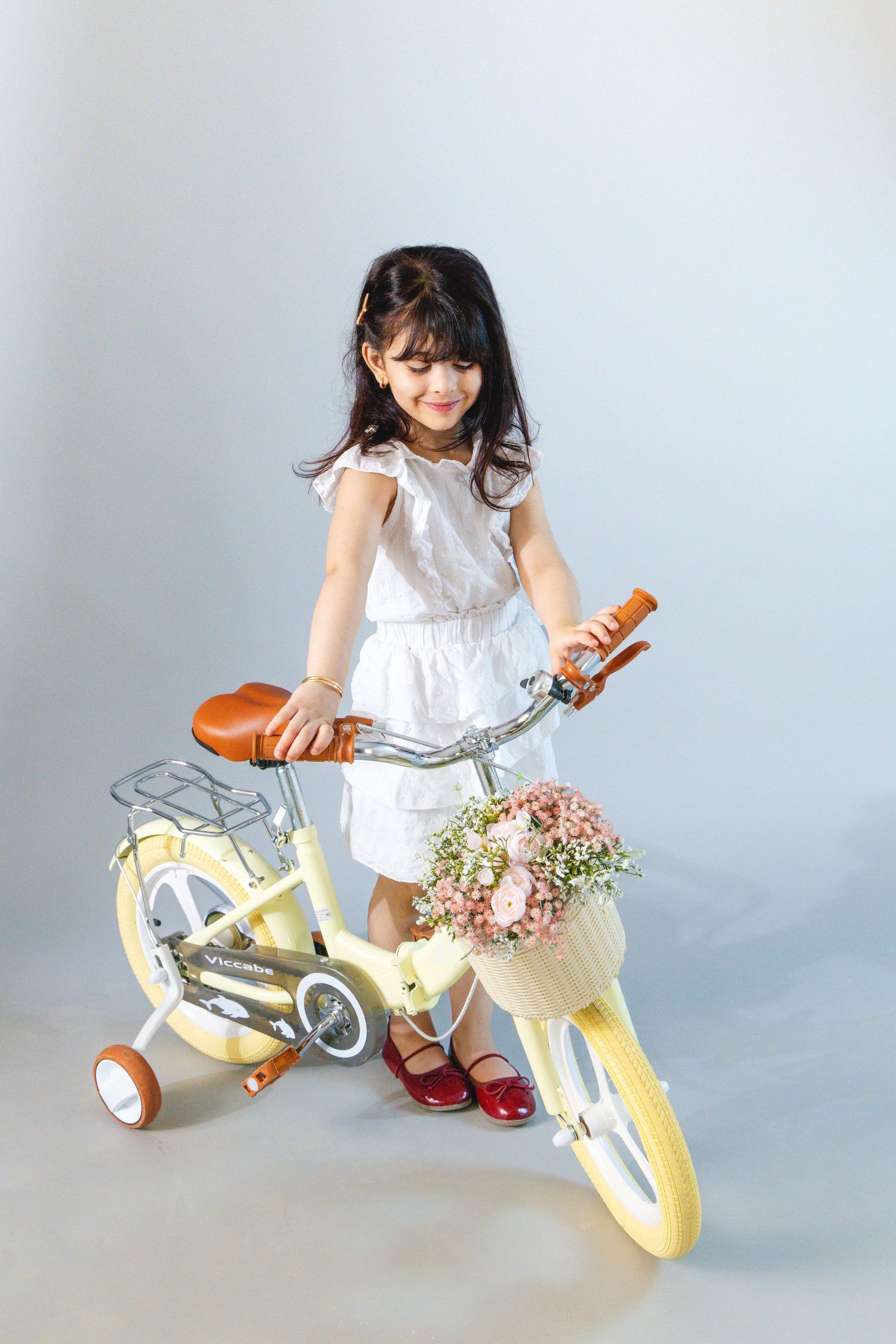 Young girl in white dress poses with yellow children's bicycle featuring training wheels and flower basket.