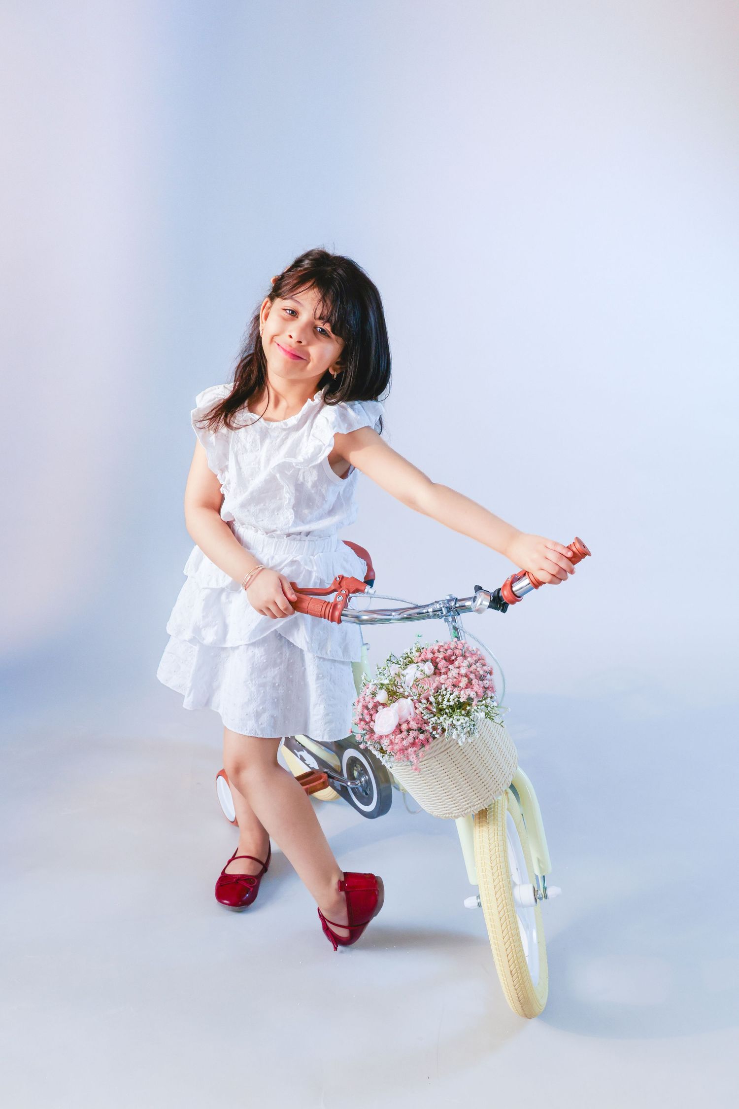 Young girl in white dress and red shoes posing with a vintage tricycle featuring a flower basket.