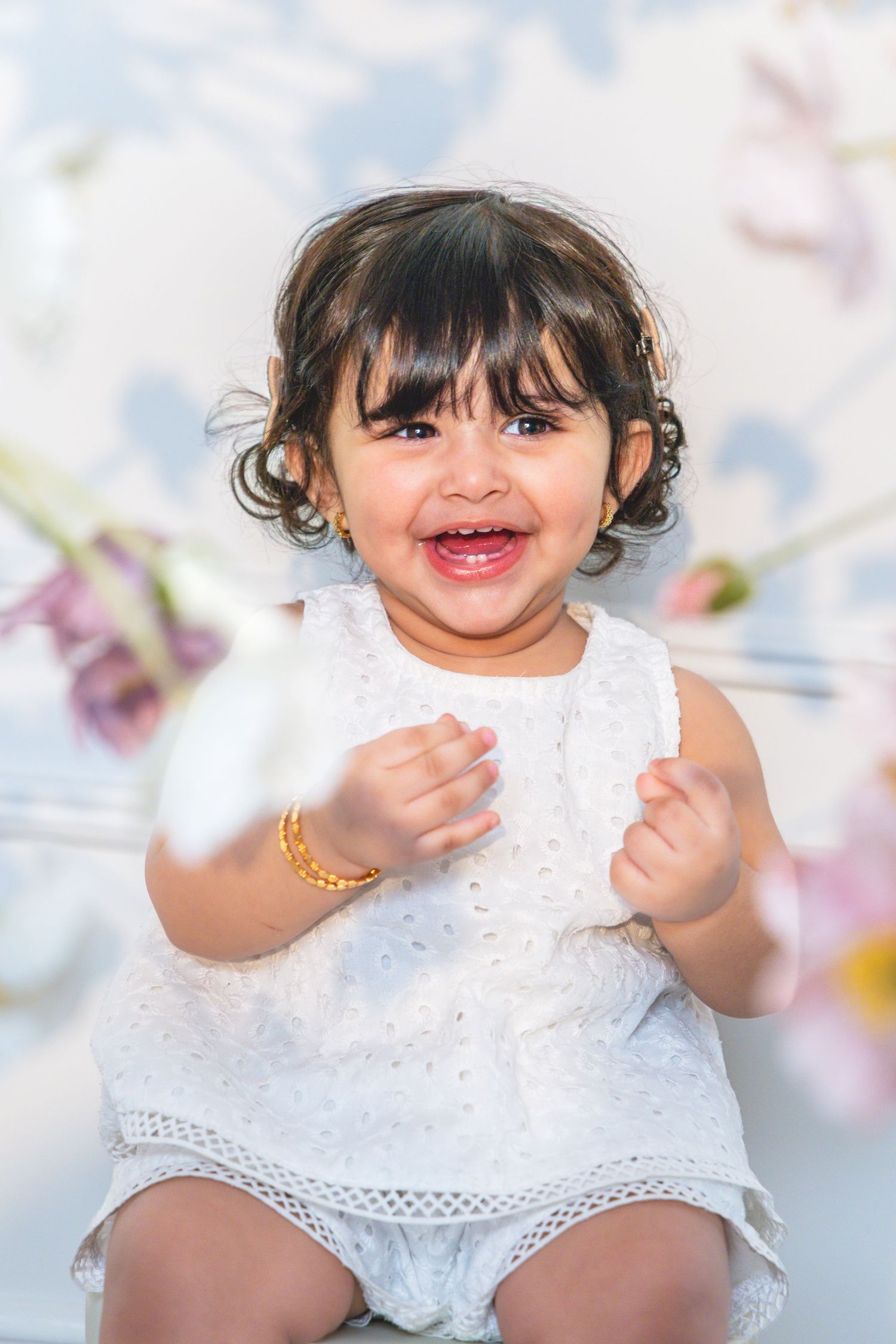 Adorable baby girl in white dress smiling joyfully, wearing gold bracelet, with floral background.