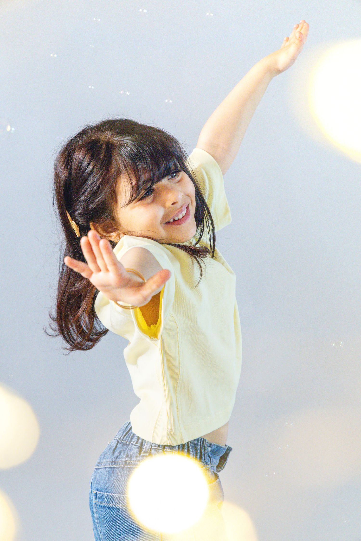 A joyful young girl in a yellow shirt and jeans dances playfully with arms raised, surrounded by bokeh lights.