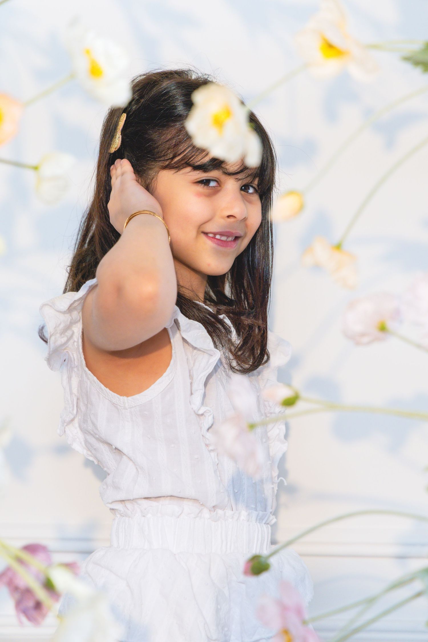 Young girl in white dress smiling among white flowers, holding a bloom near her face against a floral backdrop.
