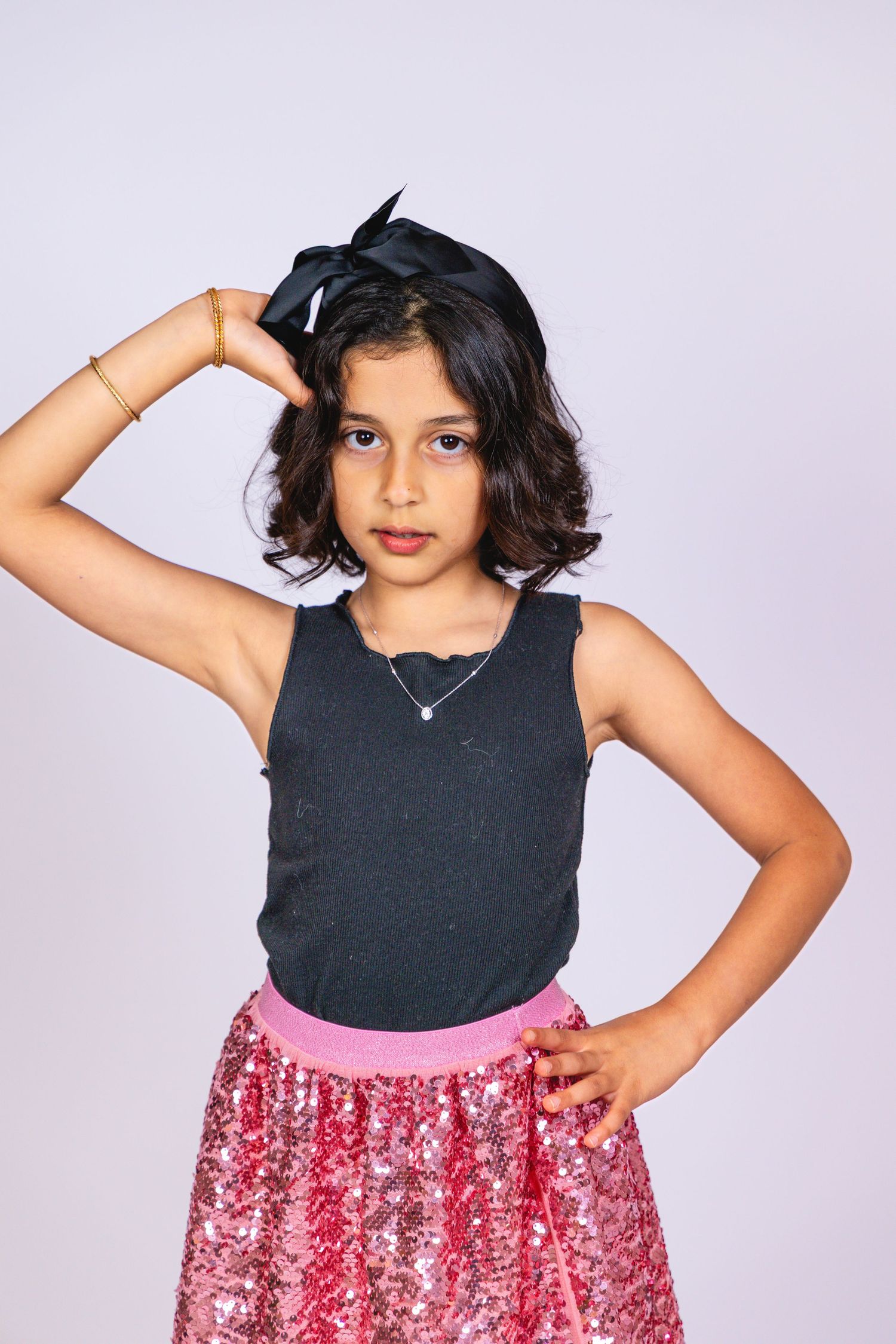 Young girl in black tank top and pink sequin skirt poses confidently with hand on hip and bow headband.