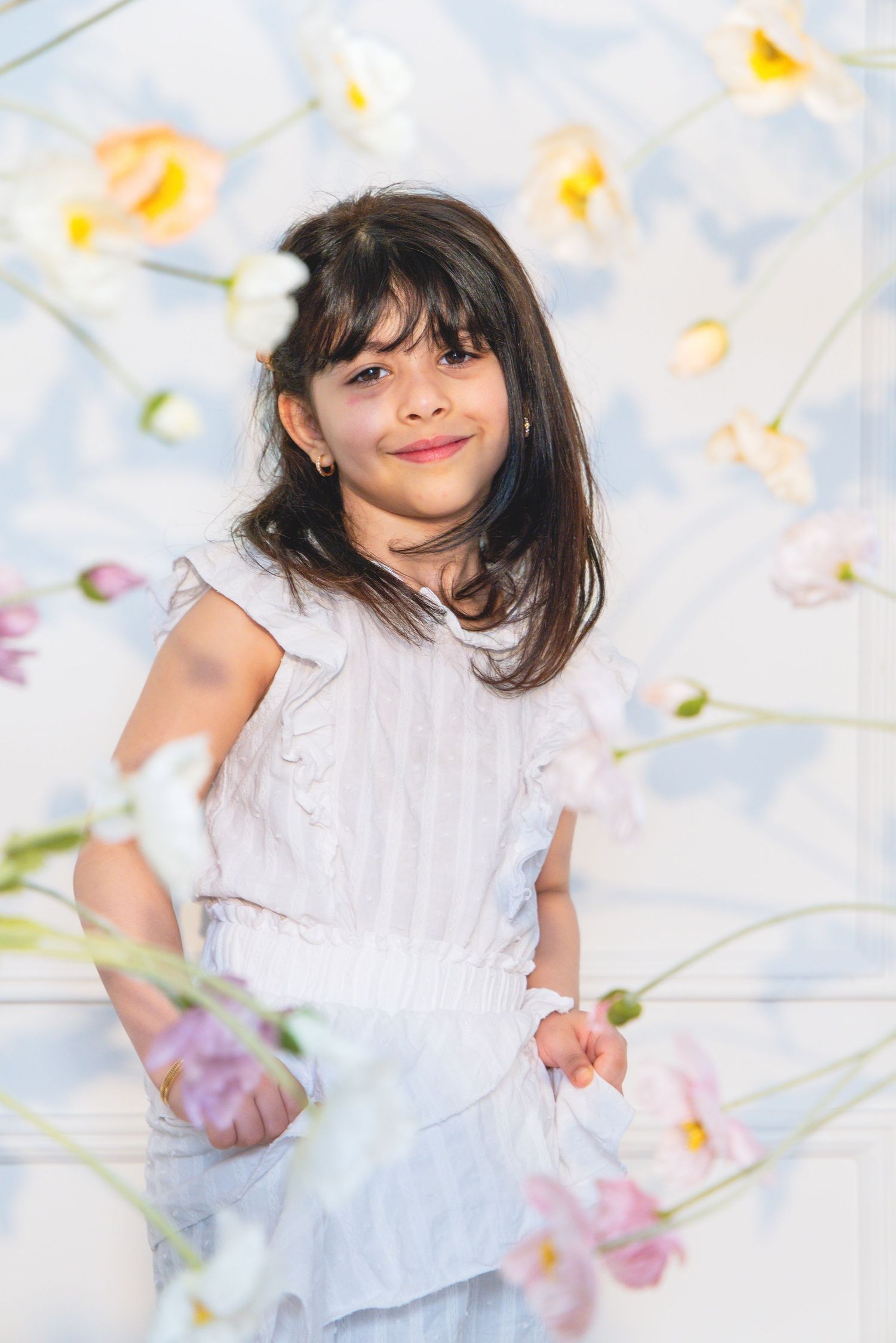 Young girl in white dress smiling among soft pastel flowers in a bright, airy floral setting.