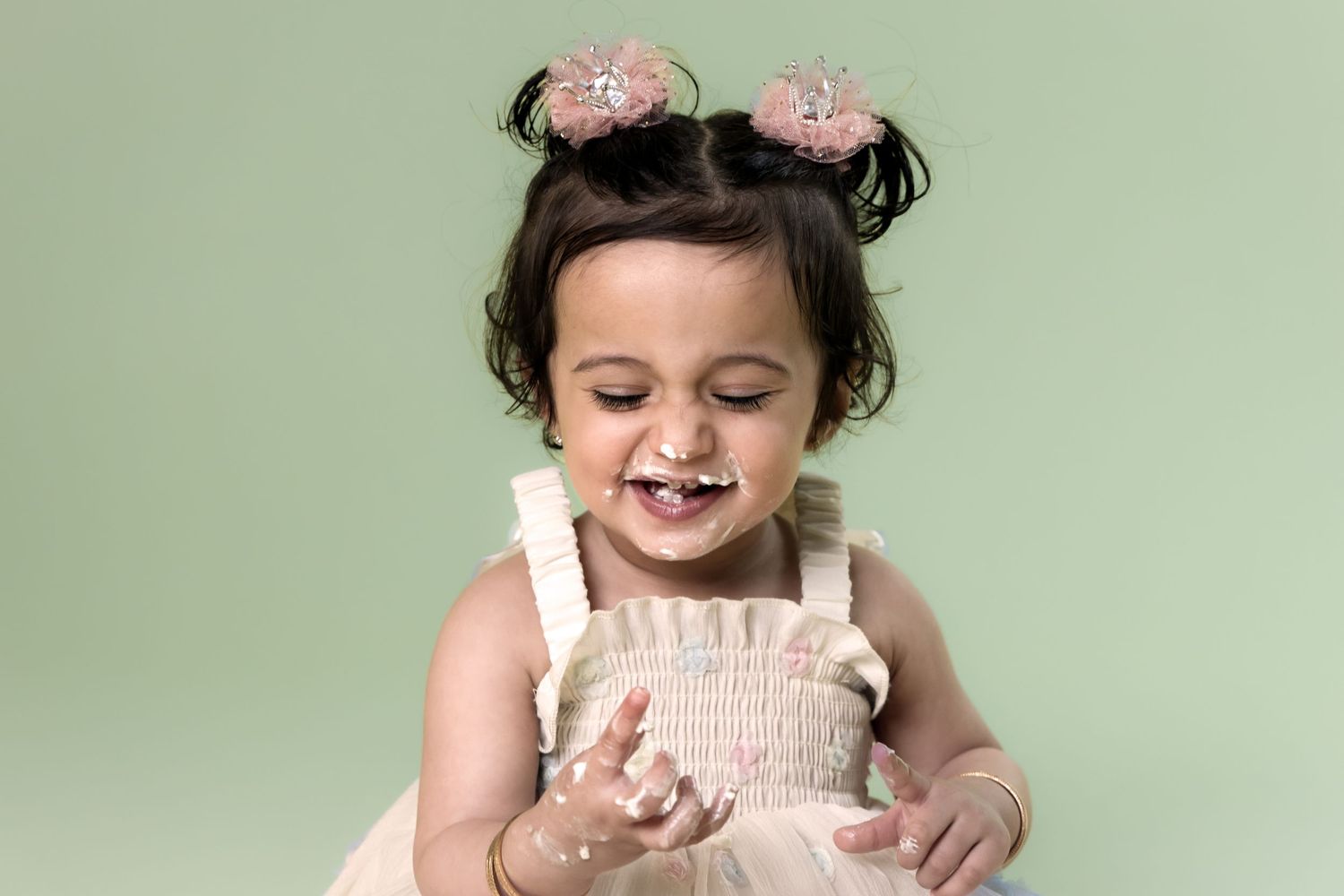Happy toddler girl with pigtails and pink bows smiling during cake smash on green background.