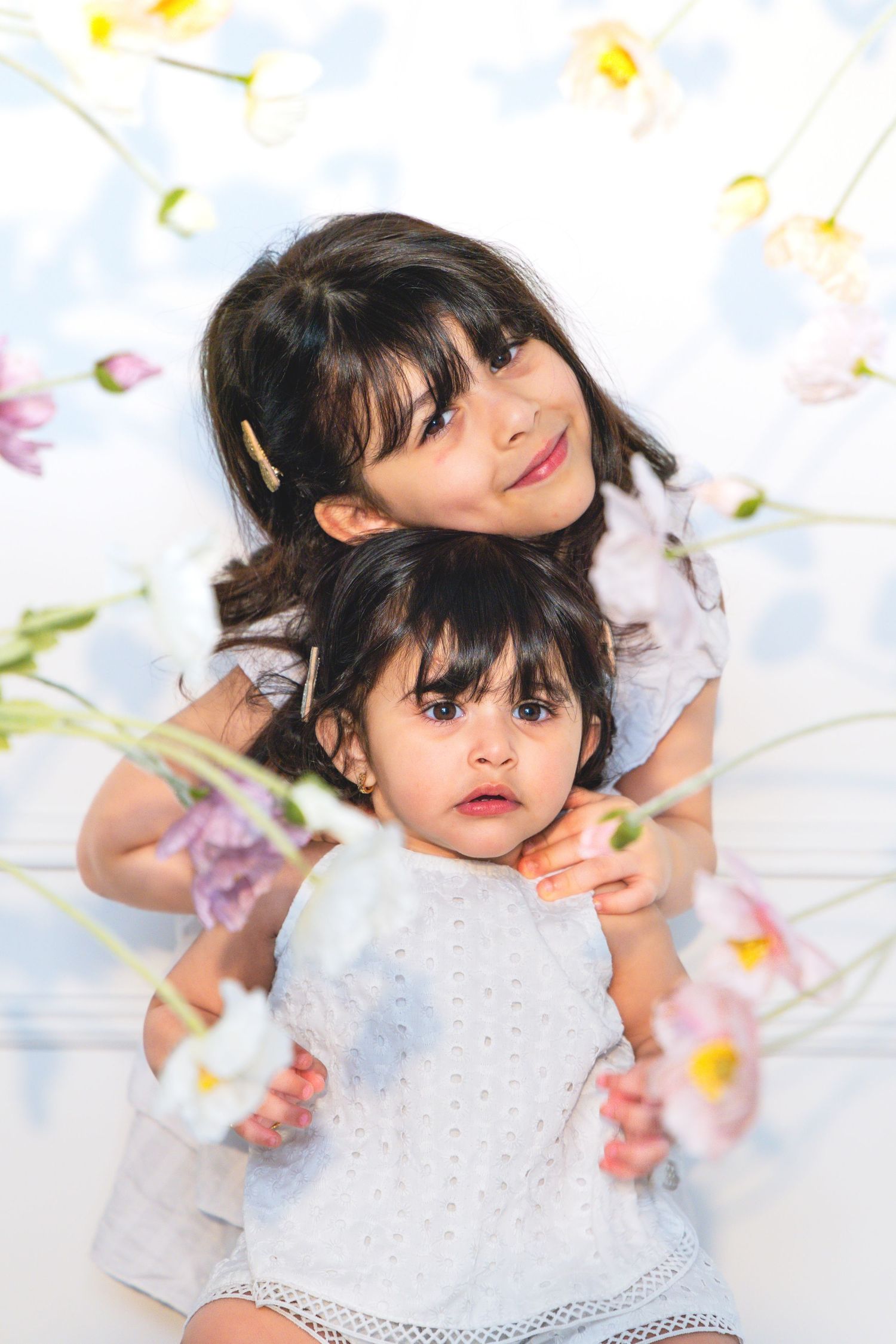 Two young sisters in white dresses pose together surrounded by delicate pink and white flowers against a light background.