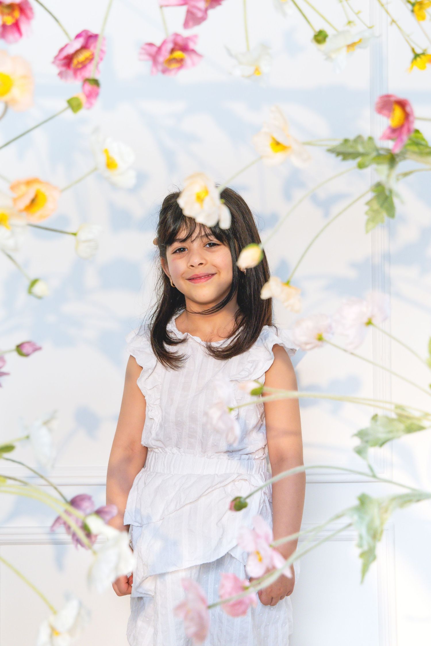 Young girl in white dress smiling among hanging spring flowers in a bright, floral studio setting.