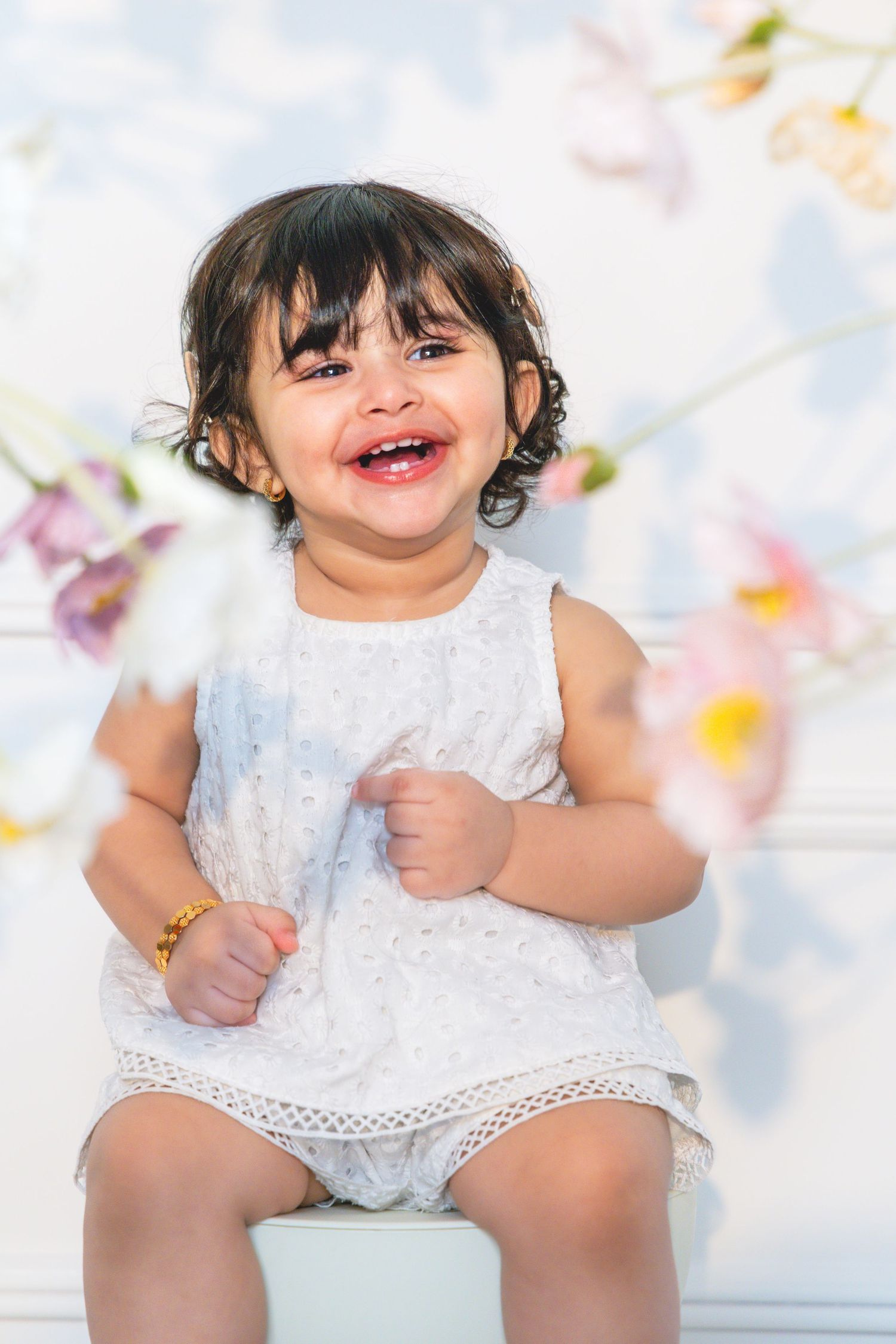 Smiling toddler girl in white dress sitting among flowers, wearing gold bracelet, with dark curly hair.