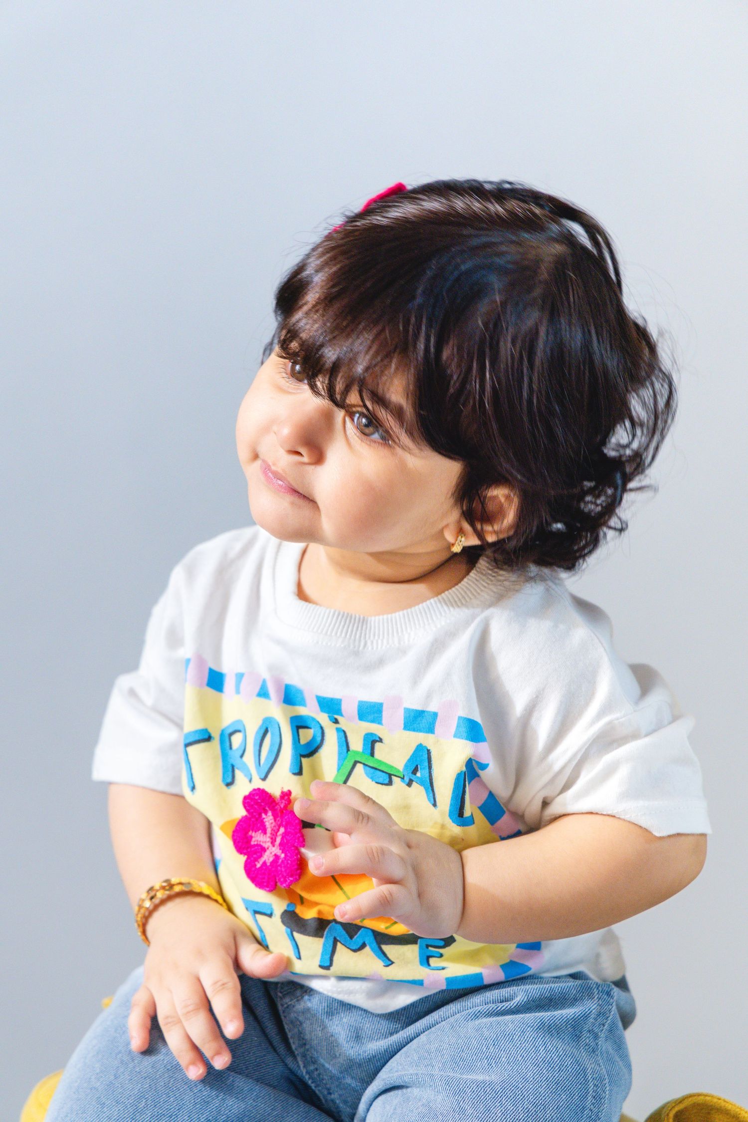 Cute toddler girl with dark curly hair wearing a white Tropical Time t-shirt and jeans, sitting against a gray background.
