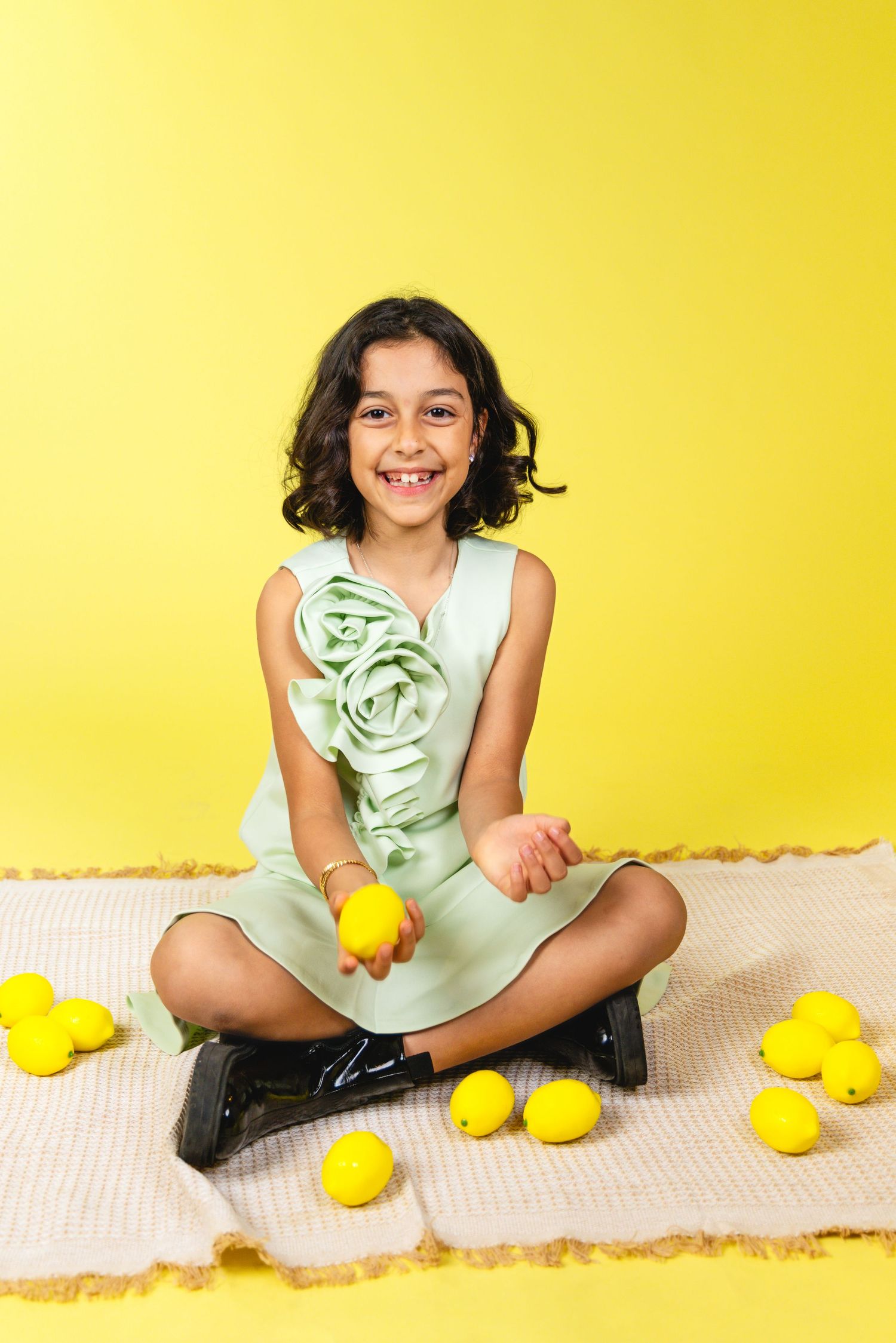 Smiling girl in mint dress sitting cross-legged on rug with lemons on yellow background.
