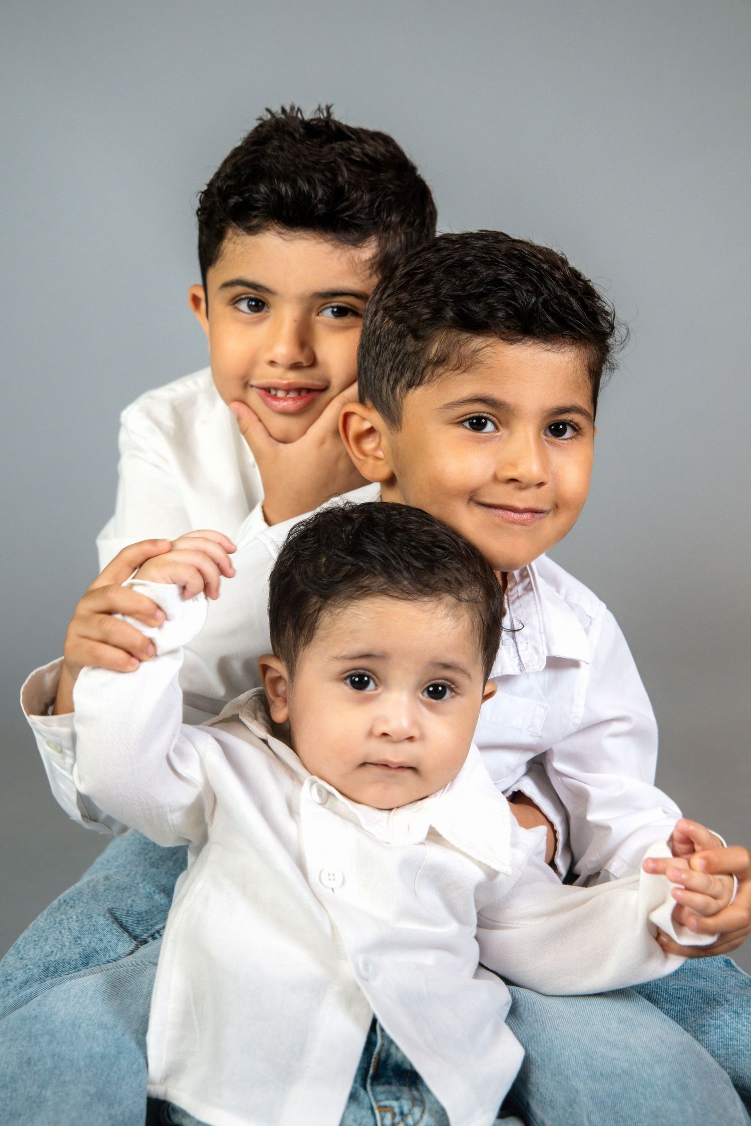 Three young brothers in white shirts pose together for a portrait against a gray background.