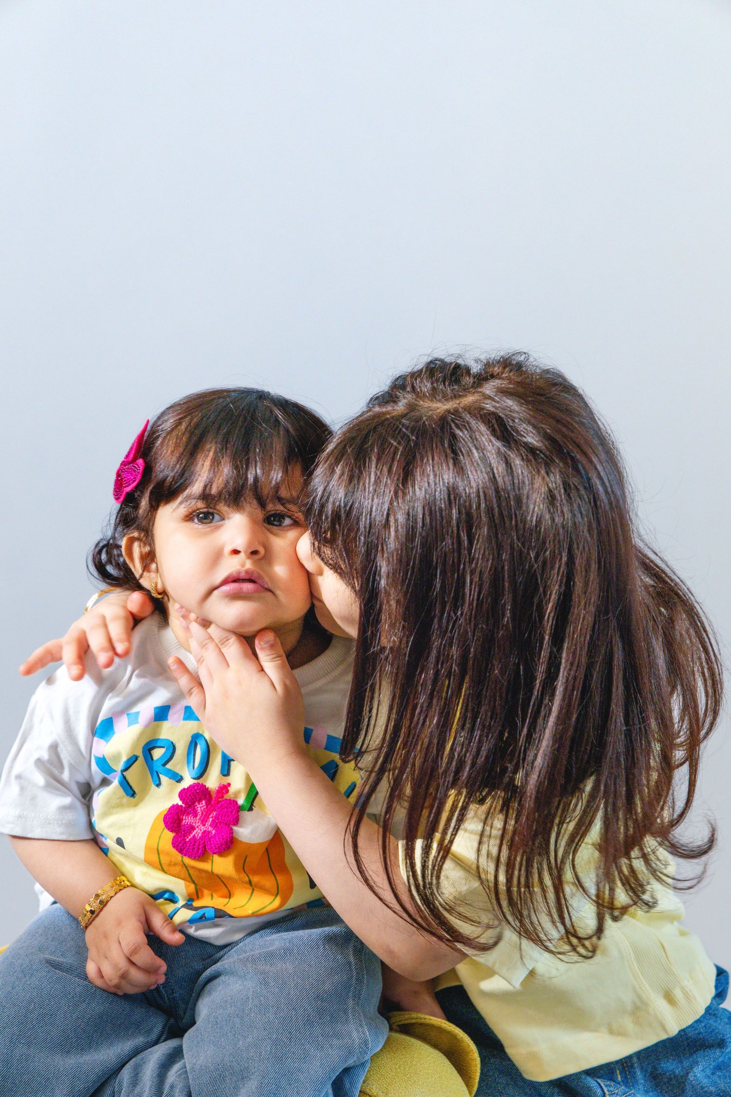 Two young girls sharing a tender moment, one whispering to the other against a light gray background.