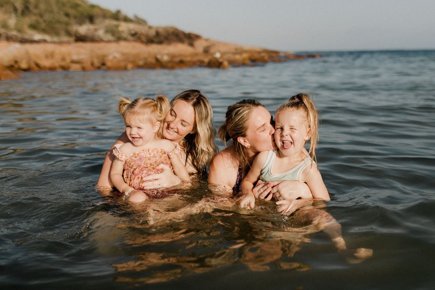 Four people enjoying a playful moment in the ocean water near a rocky coastline during golden hour.