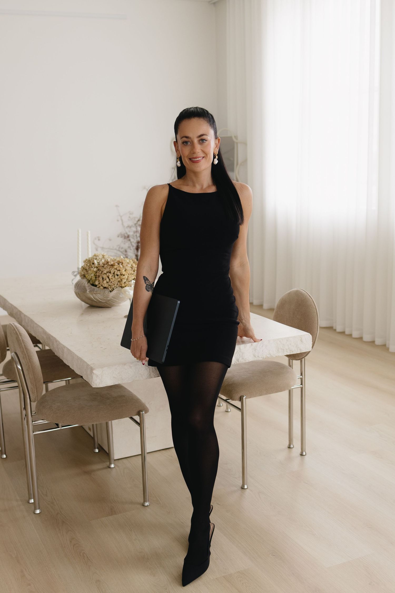 Woman in black mini dress, tights, and heels holding a clutch, standing by a marble dining table.