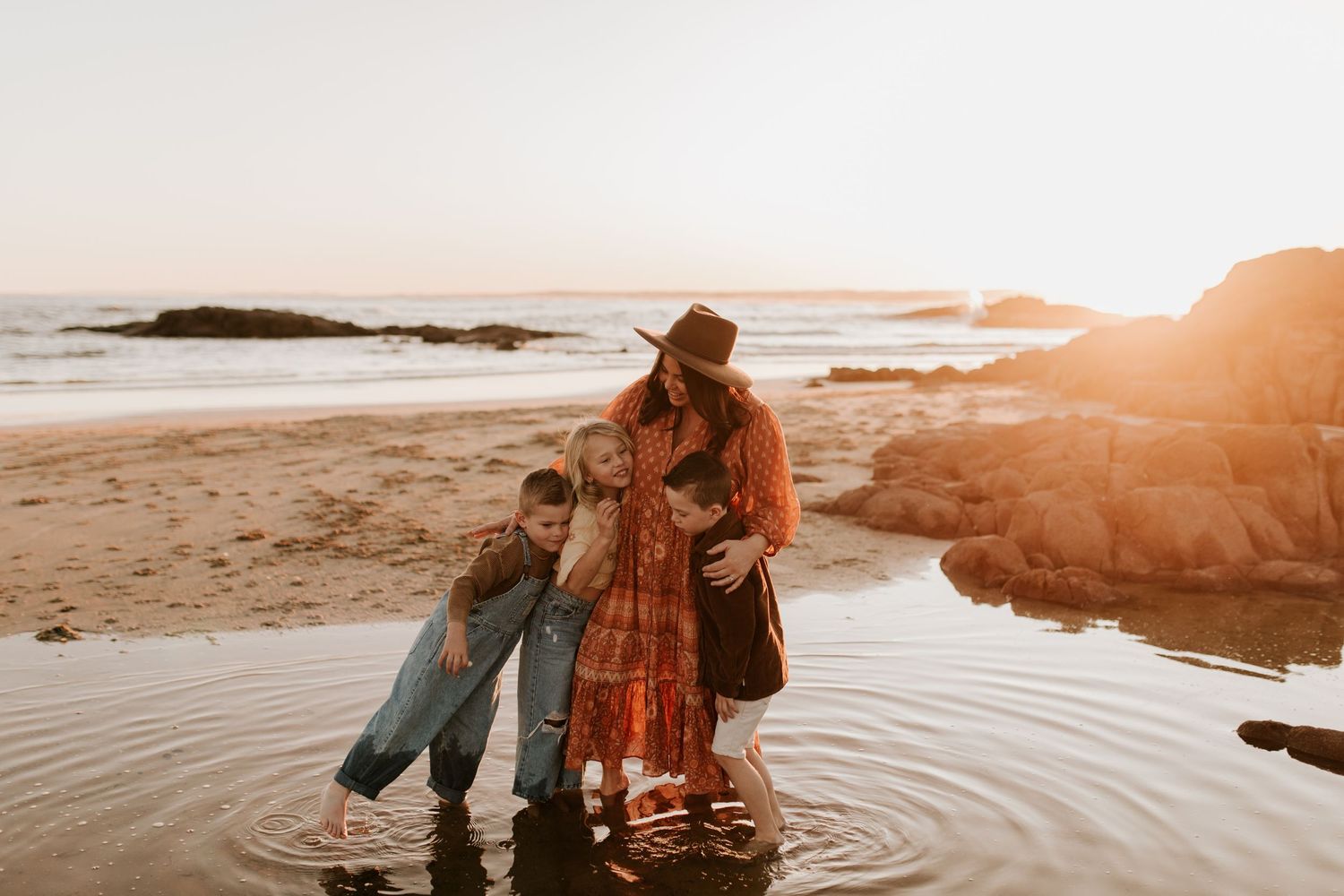 A warm sunset scene of figures splashing in shallow water at a rocky beach.