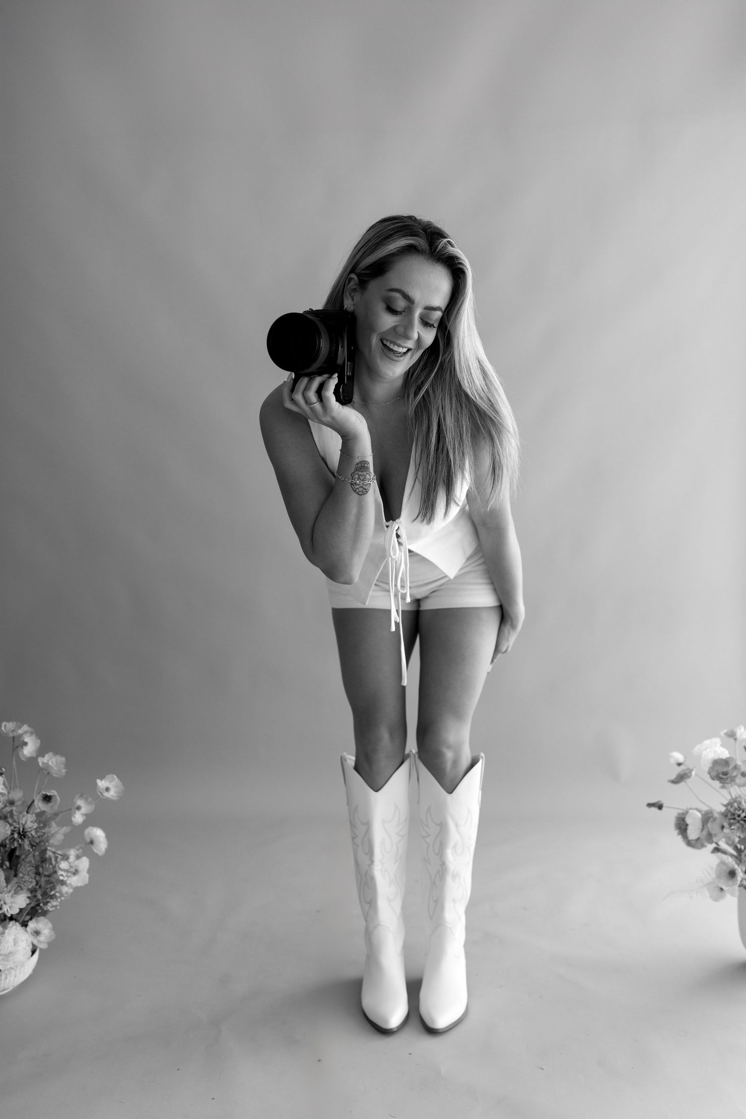 A photographer in white boots and shorts poses playfully with camera equipment in a studio setting.