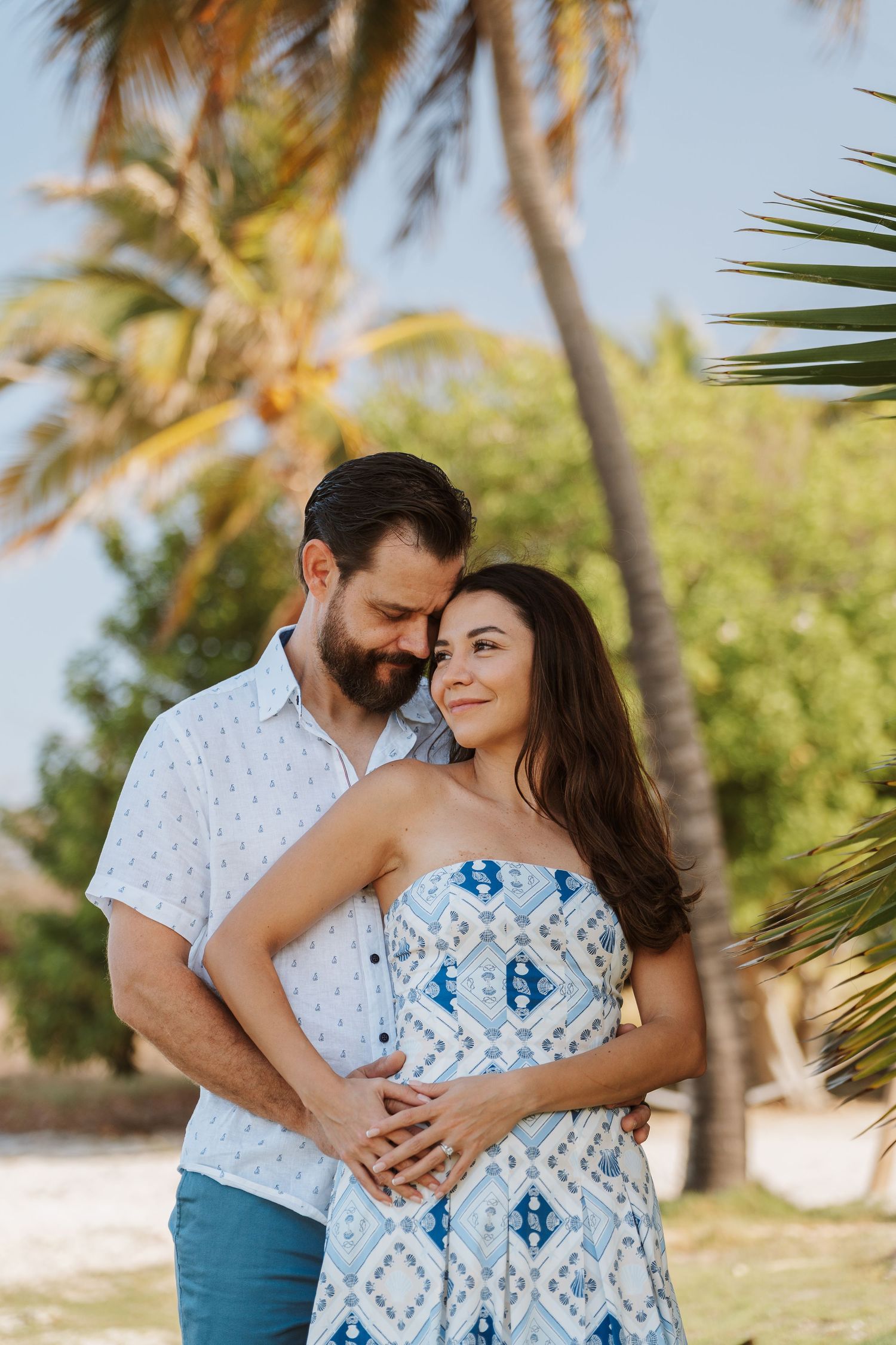 Smiling couple embracing in a tropical beach setting with palm trees, she wears a blue patterned strapless dress and he a white shirt.