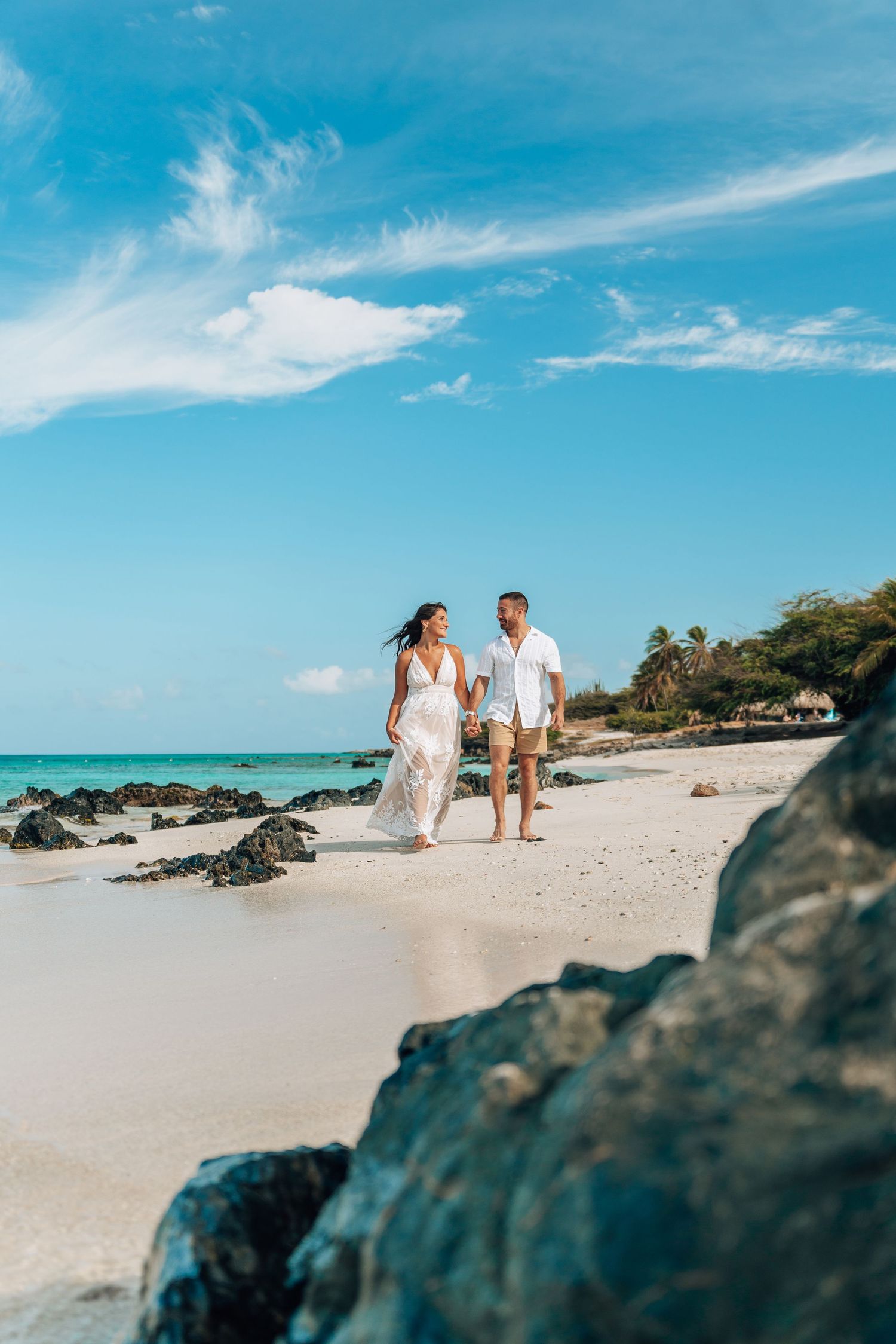 Panoramic view of a tropical beach with turquoise water, white sand, and black volcanic rocks under bright blue skies.