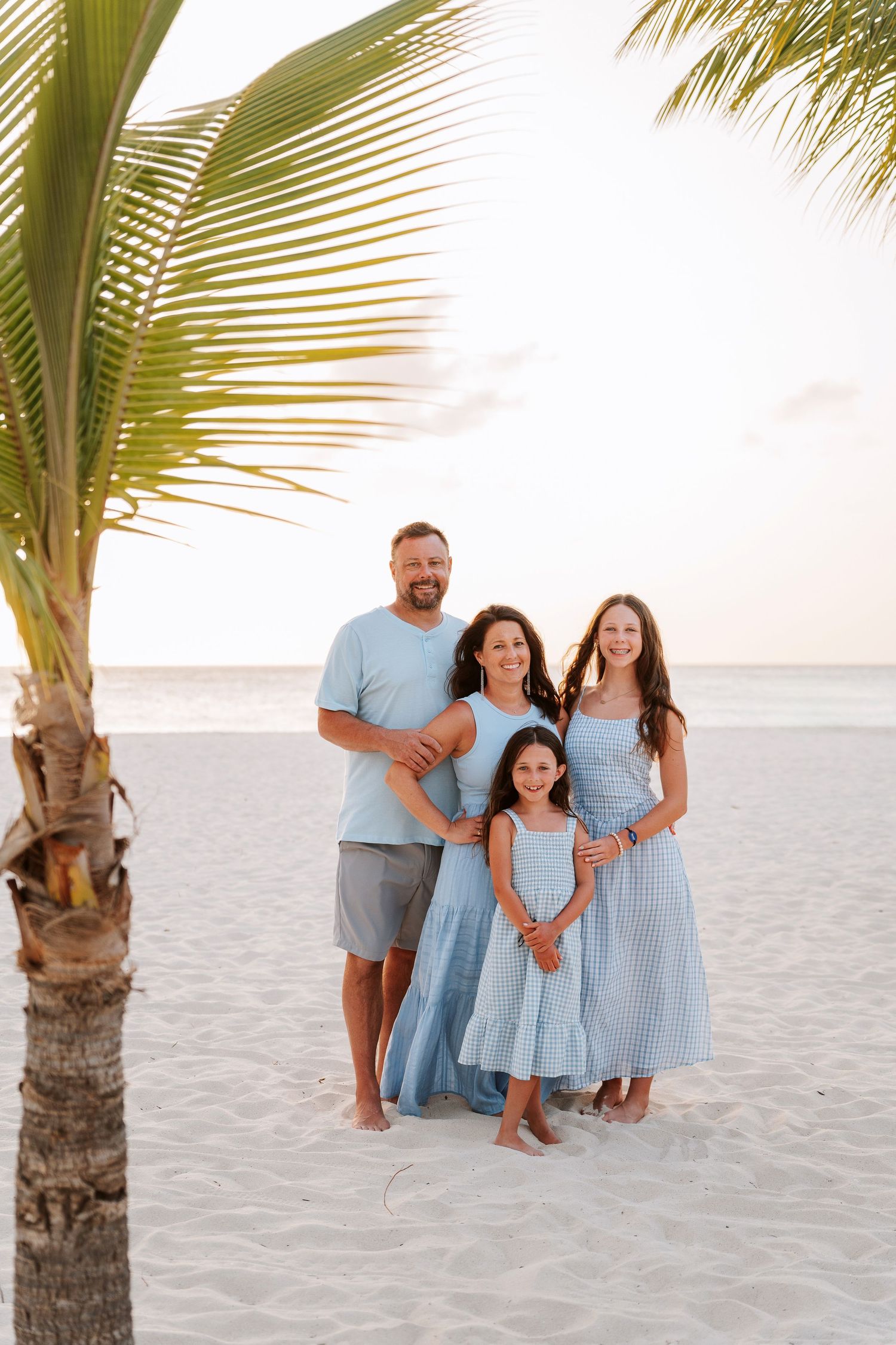 Family of four posing on white sand beach near palm tree in matching blue outfits at sunset.