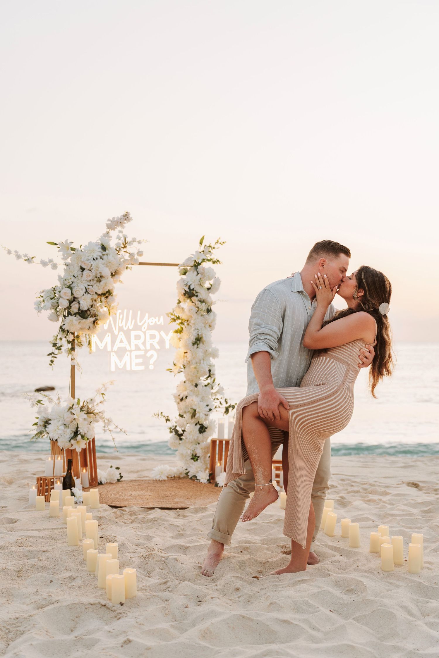 Couple kissing on beach at sunset beside a floral arch with Will You Marry Me sign, surrounded by candles.