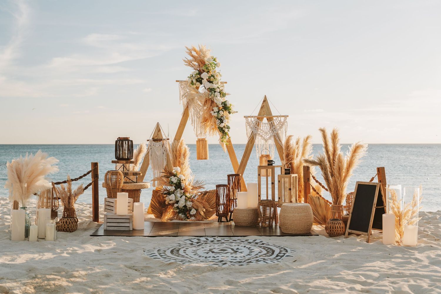 Boho beach wedding setup with geometric arches, pampas grass, lanterns, and floral decor on sandy shore at sunset.