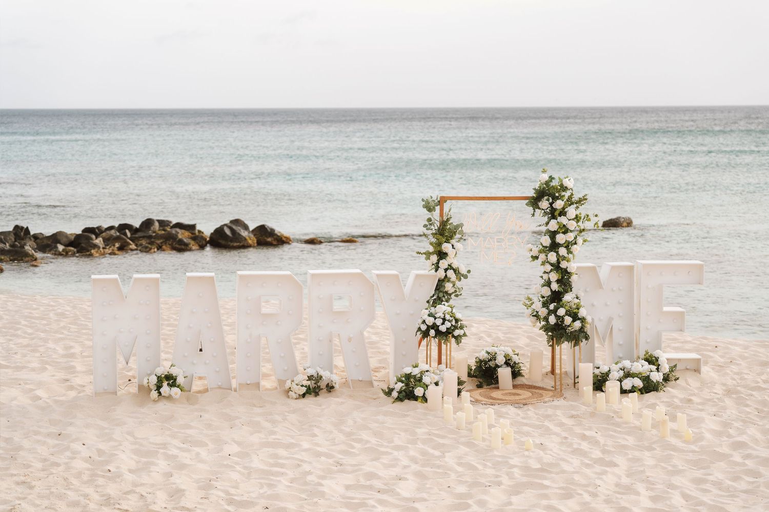 Beach wedding setup with MARRY ME letters, floral arch, candles, and white flowers on sandy shore with ocean backdrop.