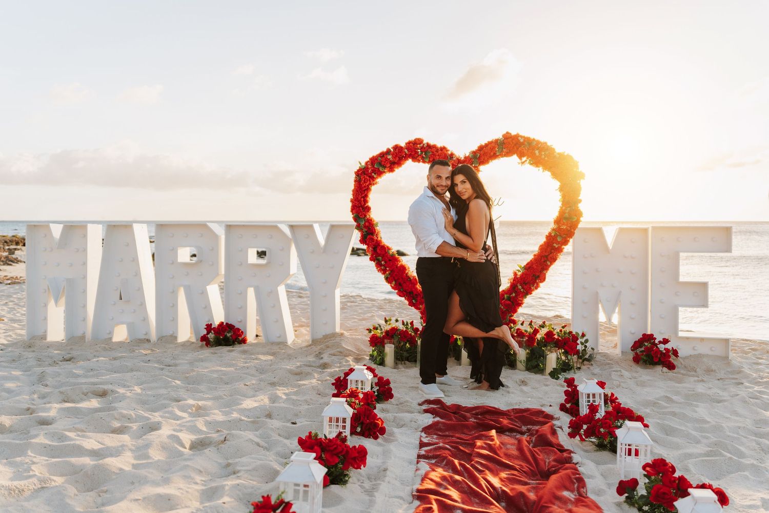 Couple embraces at romantic beach proposal with MARRY ME letters, red rose heart, and flower arrangements at sunset.