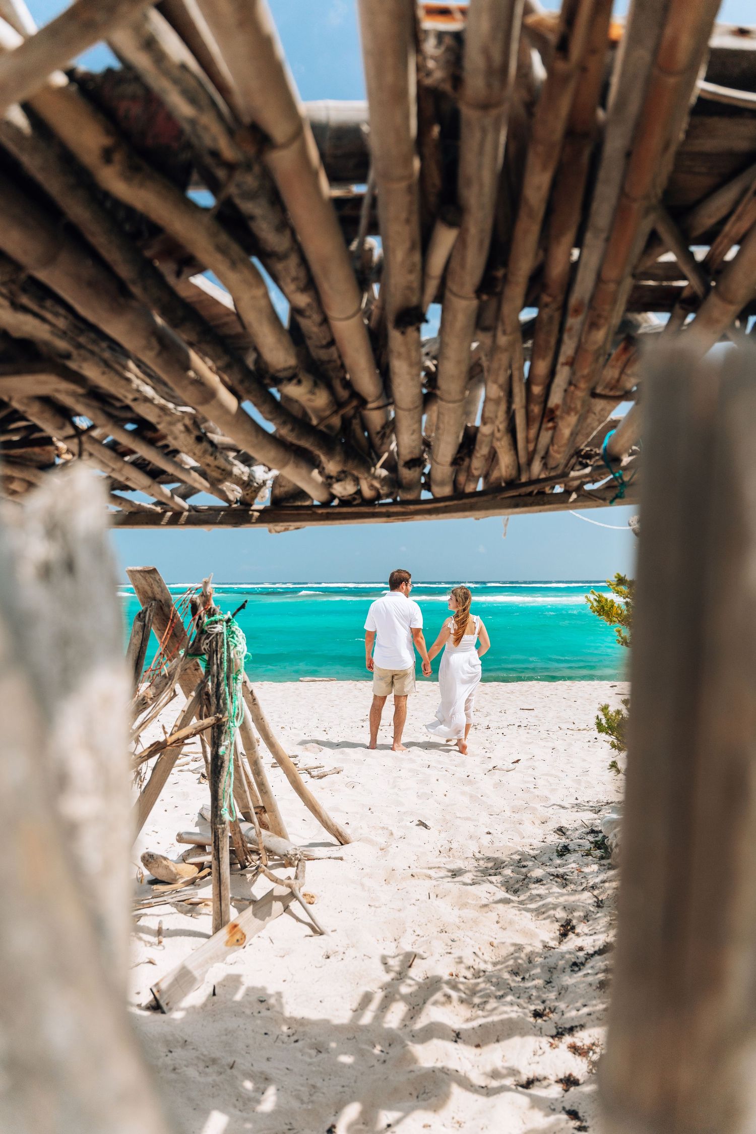 Couple walking on white sandy beach viewed through rustic driftwood structure with turquoise ocean waters in background.