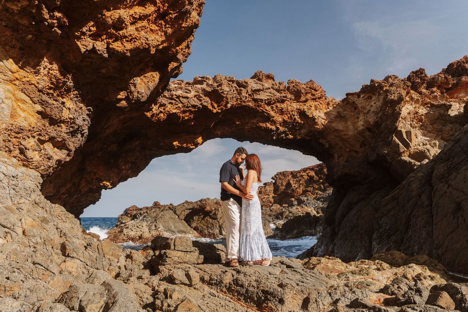 Two people embrace under a natural rock arch formation in a desert landscape at sunset.