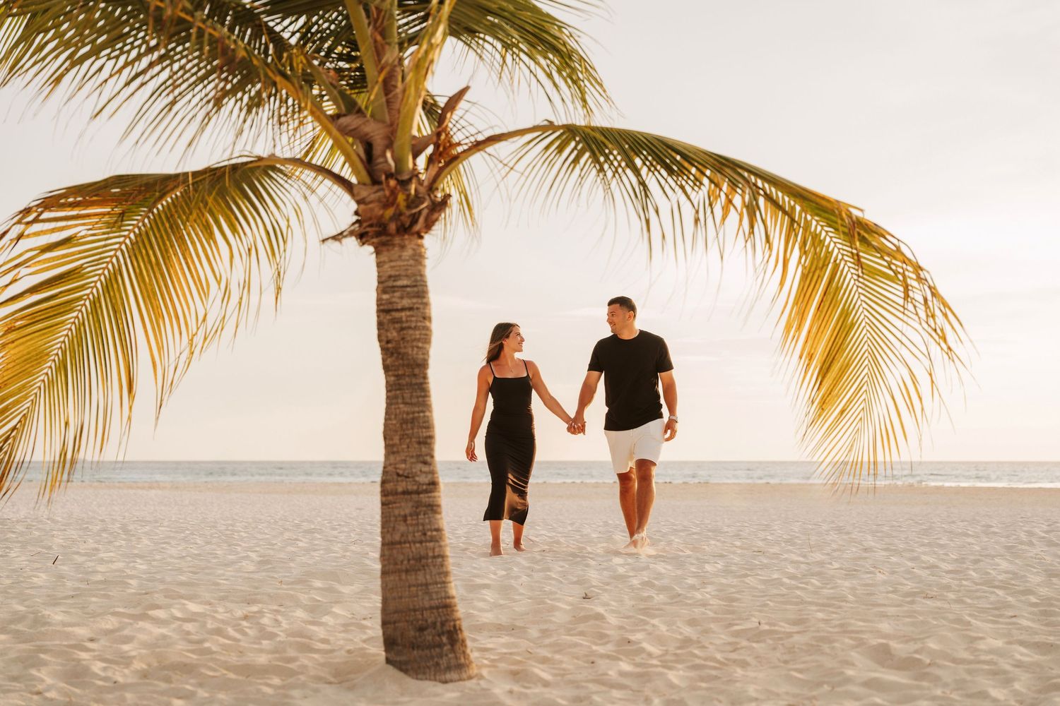 Two figures walk along a tropical beach with palm trees and soft sand during golden hour.
