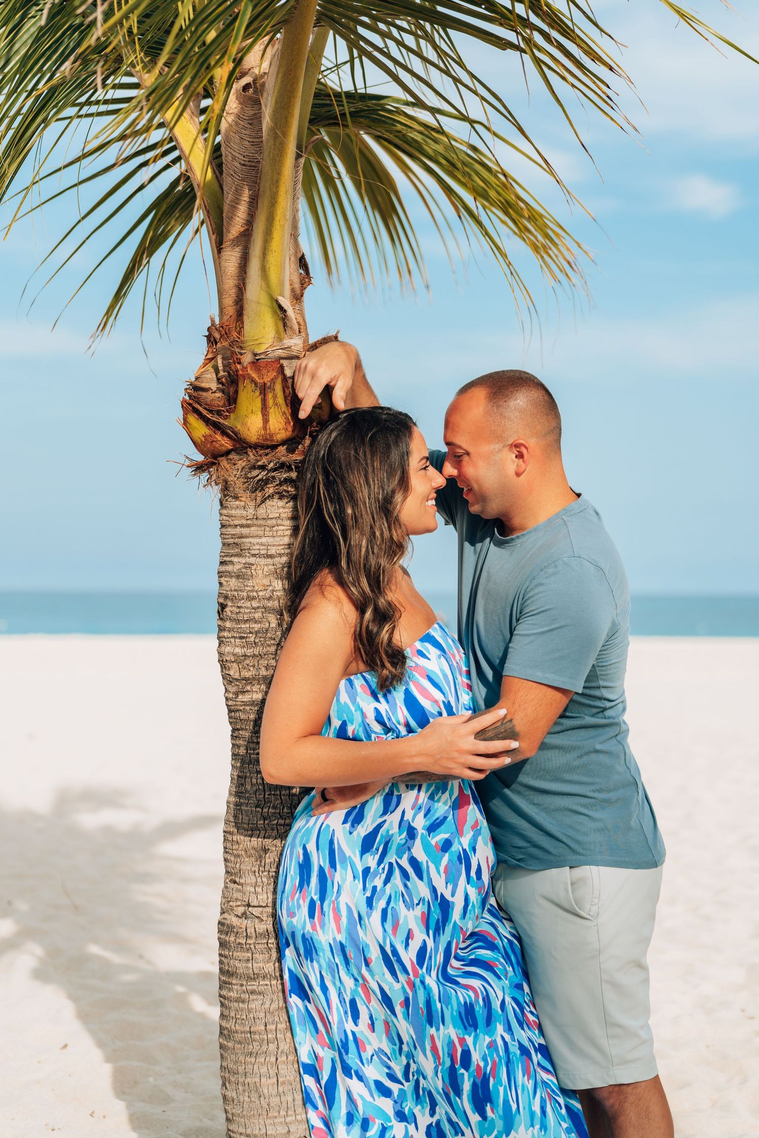 Couple sharing a romantic moment under a palm tree on a white sand beach, woman in blue floral dress.