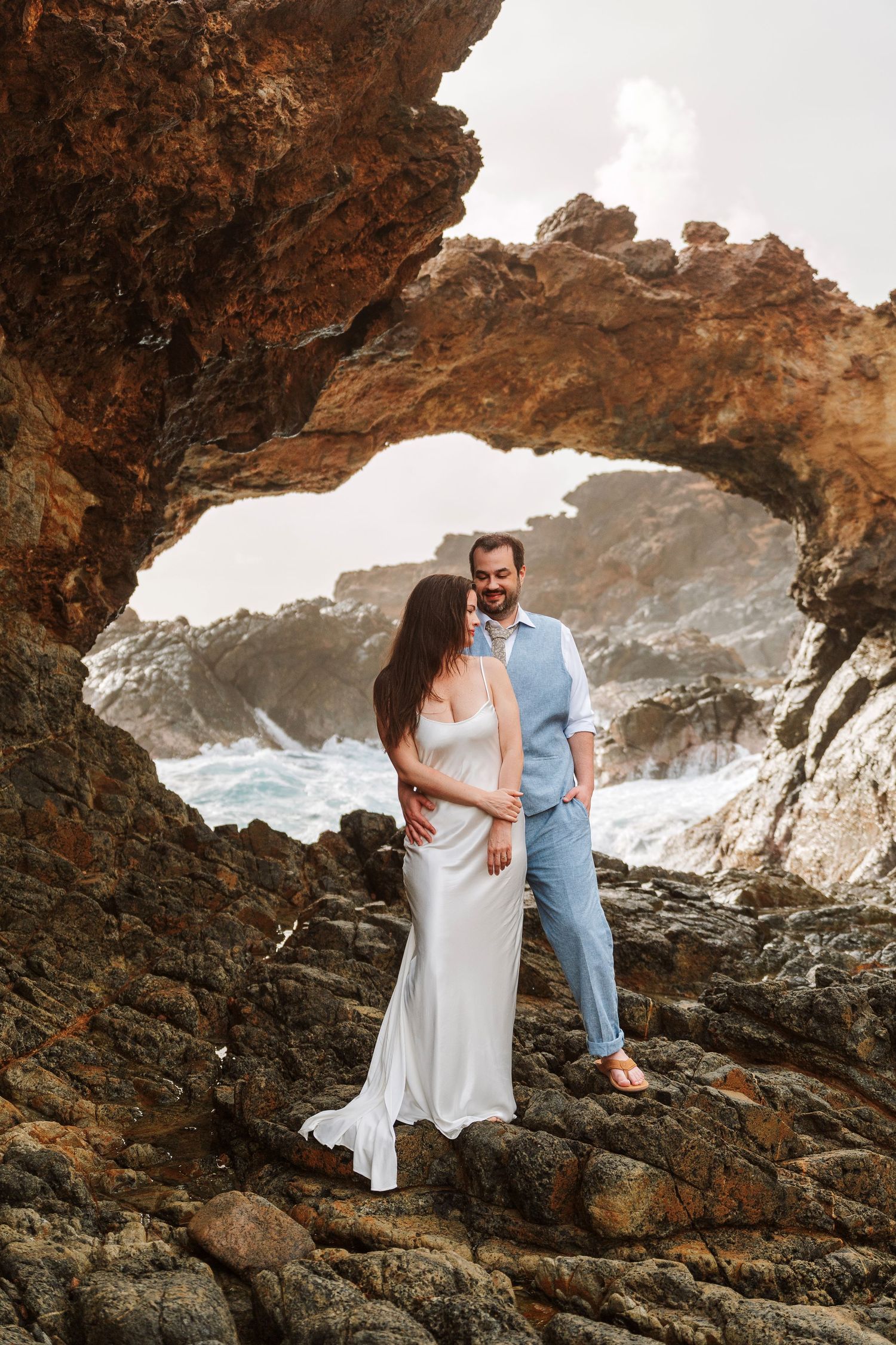 Couple in wedding attire embracing under a natural rock arch on a rocky coastline with ocean waves behind them.
