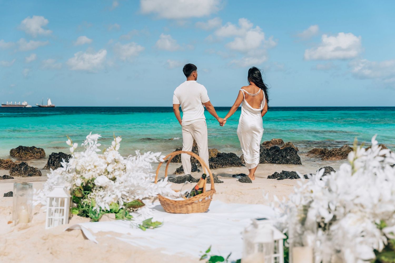A couple holds hands while walking along a pristine turquoise beach with white sand and rock formations.