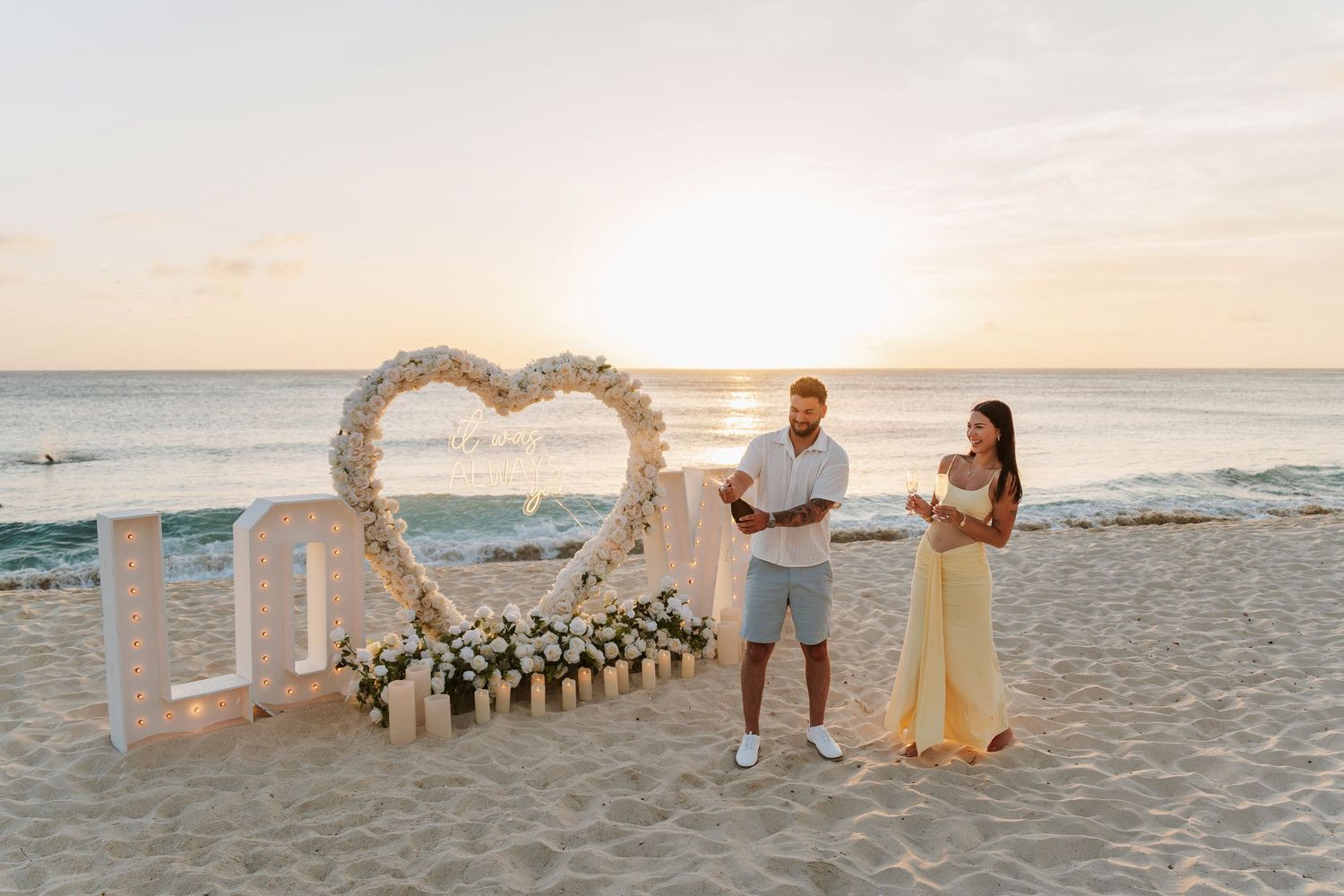 Couple stands by floral heart arch and LOVE letters on a sandy beach at sunset during a romantic proposal.