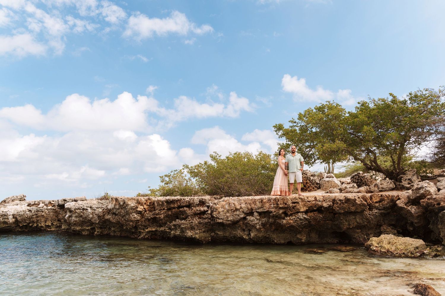 Couple standing on rocky coastal cliffs surrounded by turquoise water and lush green trees under a bright blue sky.