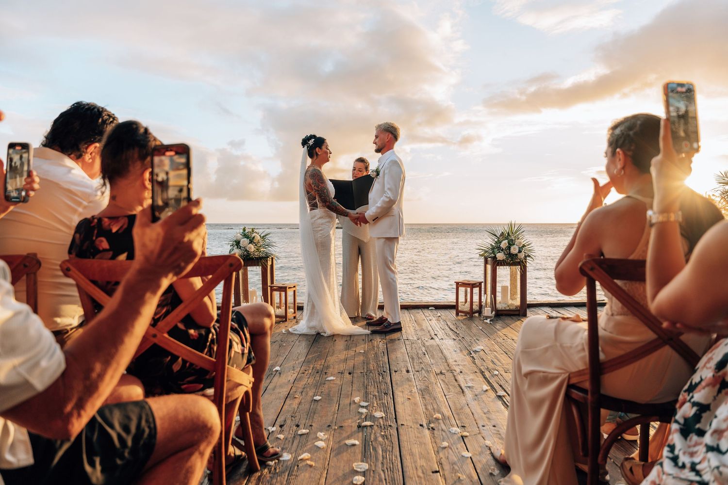Couple exchanges vows at a romantic beachside wedding ceremony on a wooden deck at sunset with guests watching.